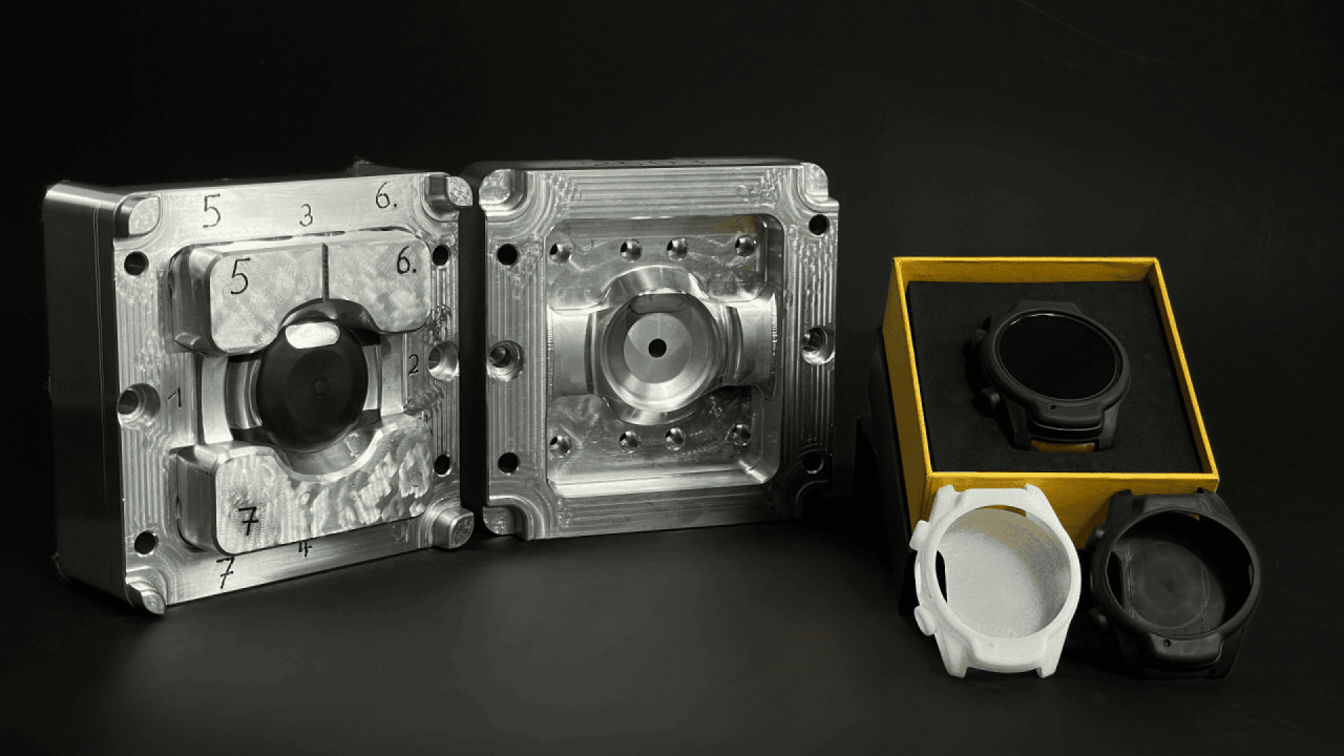 Two metal molds are displayed side by side, with a black and a white watch case in front of them, and a finished black watch in a yellow box on the right, all set against a black background.