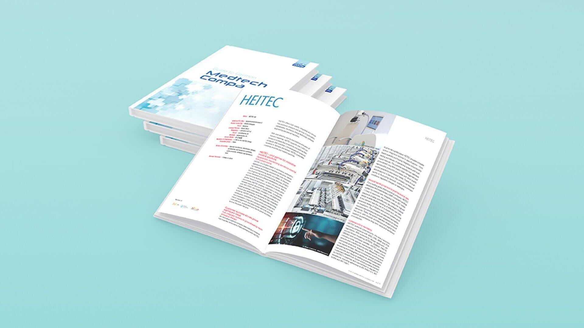 A stack of books titled Medtech Comp lies on a light blue surface, with one book open to reveal pages of text, pictures of industrial equipment and the inside of a factory.