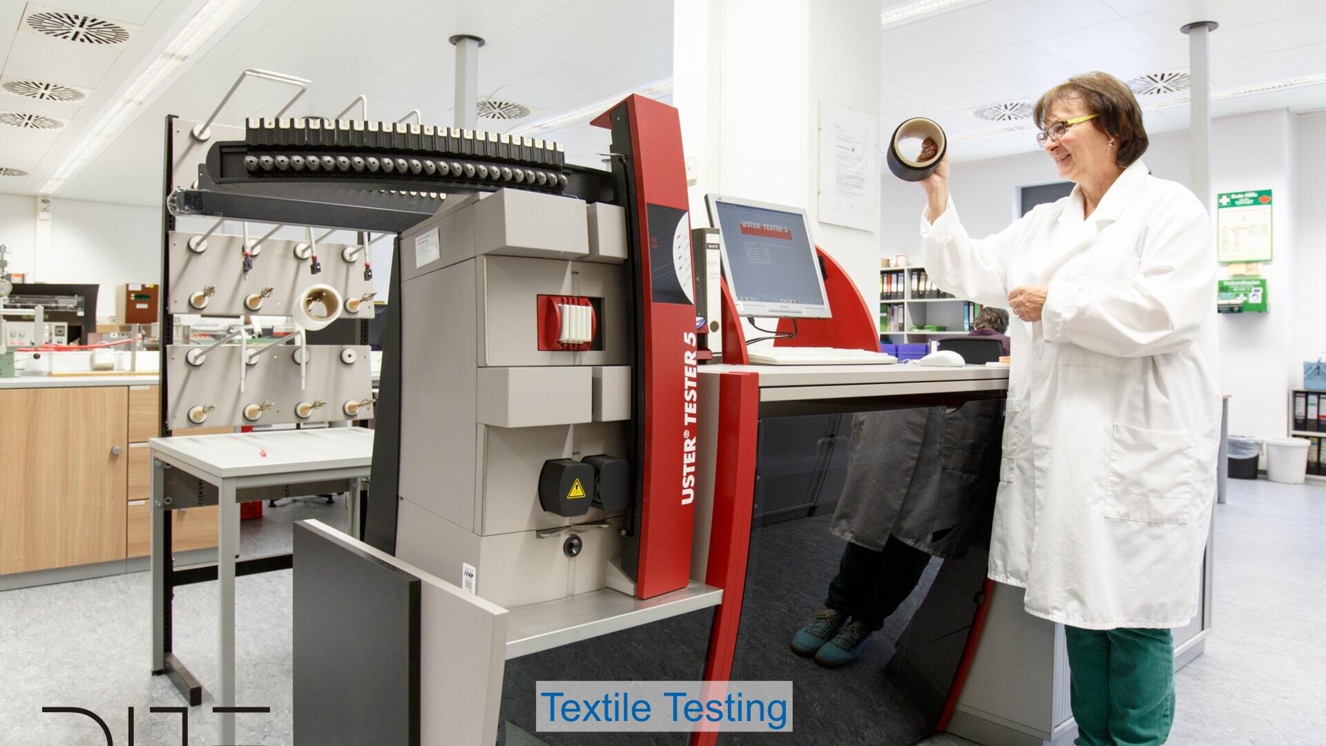 A woman in a lab coat smiles while holding a spool of thread in a laboratory. She stands beside a textile testing machine with a computer and several thread samples in the background.