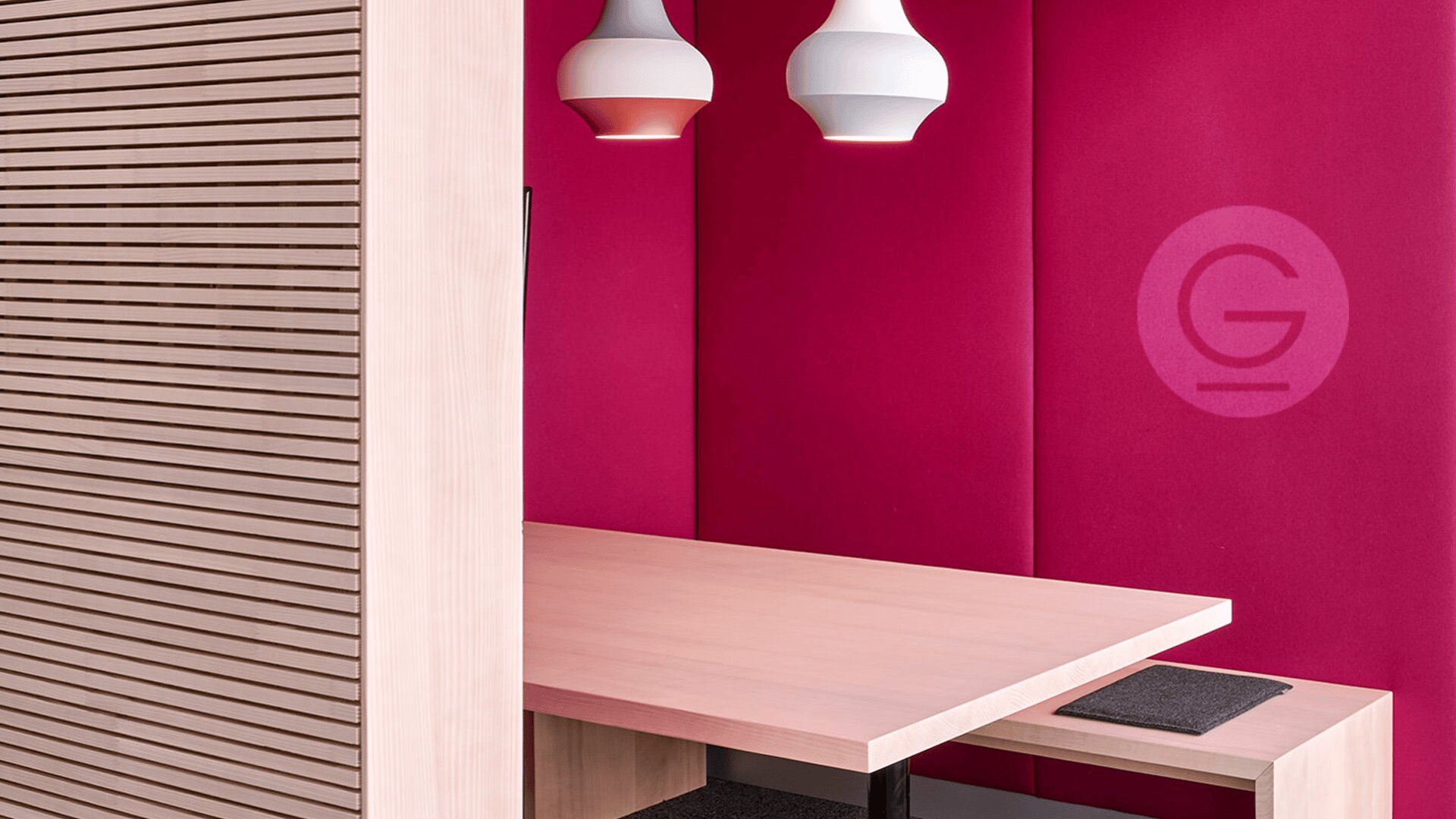 Moderne Besprechungsecke mit Holzbank und -tisch, zwei weißen Pendelleuchten und magentafarbenen gepolsterten Wänden; links ist teilweise eine Trennwand aus Holzlatten sichtbar.