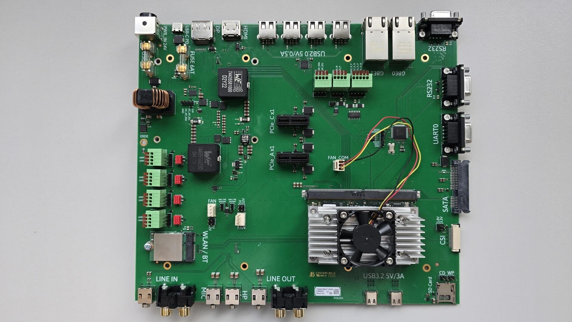 A green circuit board with various electronic components, ports, connectors, and a small cooling fan attached to a metal heat sink in the lower right section. The board is labeled with white text and symbols.