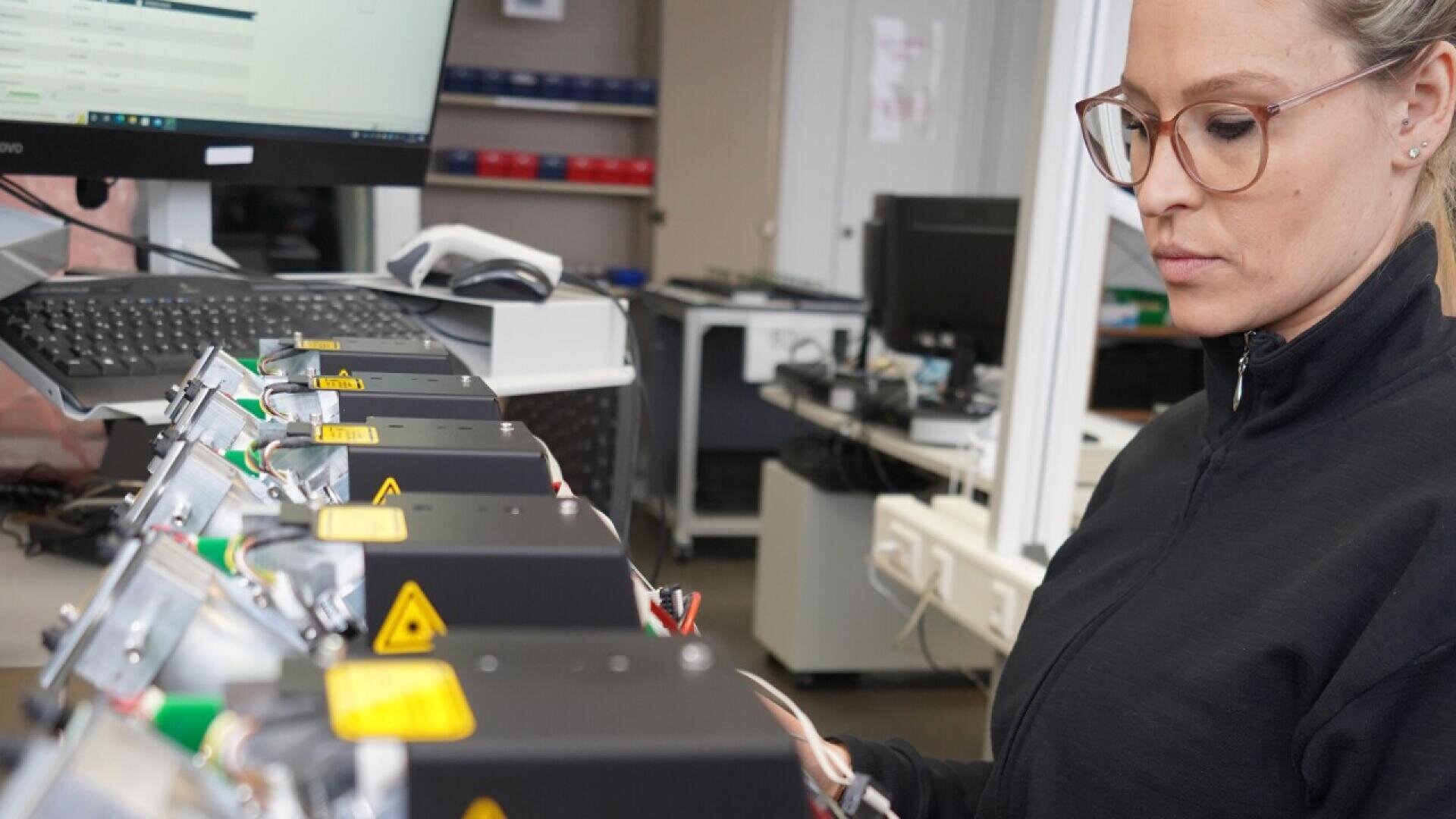 A woman wearing glasses and a black jacket inspects a row of electronic devices in an office or laboratory, with a computer monitor and office equipment in the background.