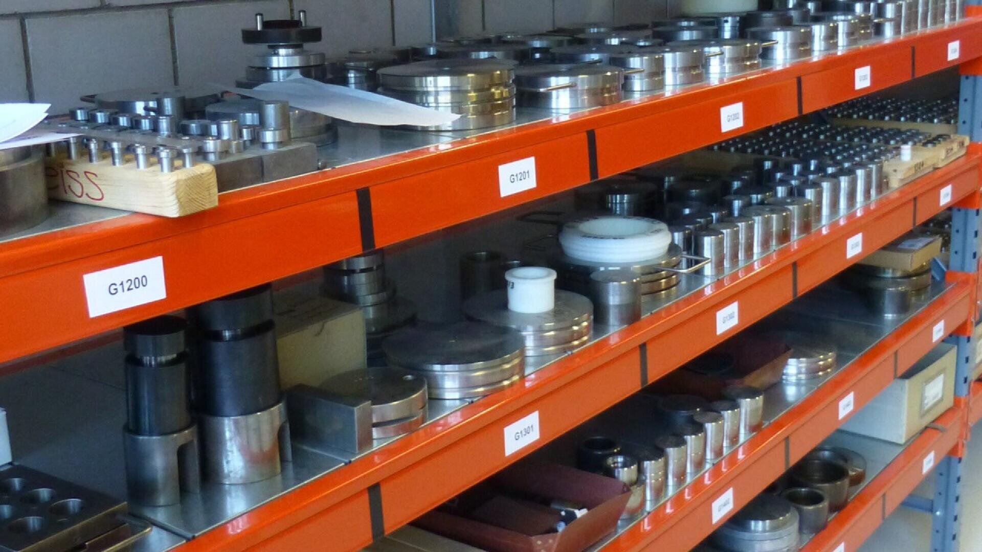 Three shelves lined with metal parts and components, arranged neatly and labeled by section. The items vary in size and shape, and the shelves are painted orange. The background shows a tiled wall.