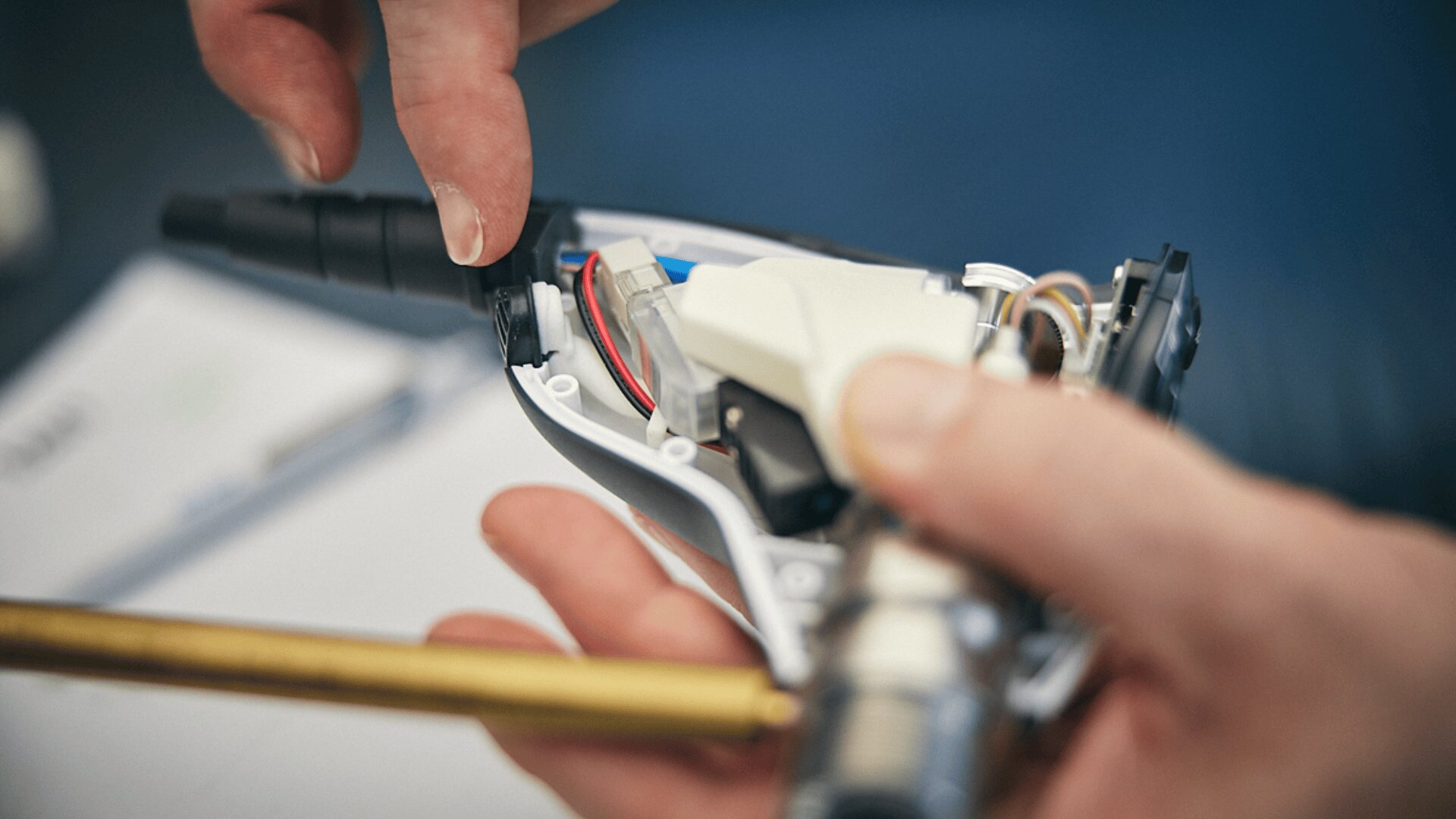 Eine Nahaufnahme von Händen, die ein elektronisches Gerät mit freiliegenden Drähten und Bauteilen zusammenbauen oder reparieren, vor einem unscharfen Hintergrund.