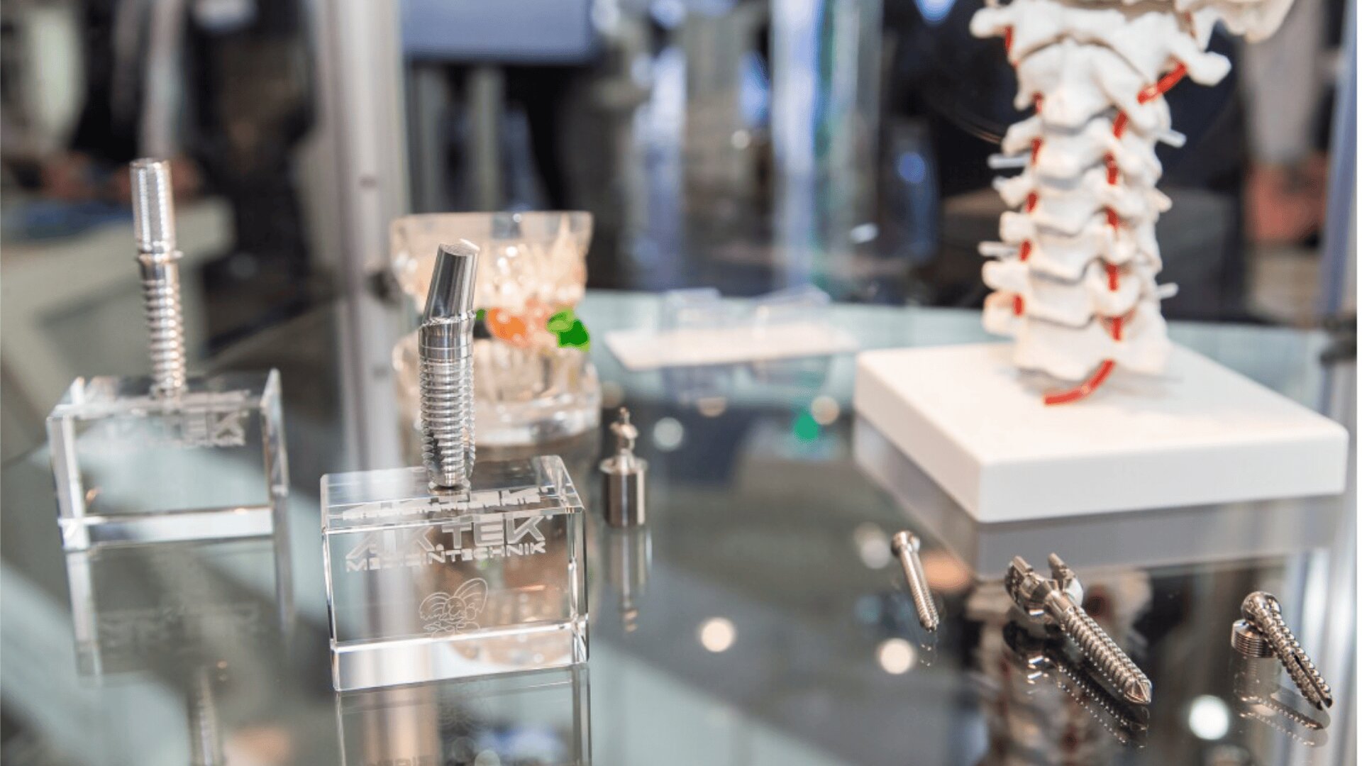 A display of medical implants, including metal spinal screws and rods, on a glass table. A model of a human spine is shown in the background, illustrating surgical procedures.
