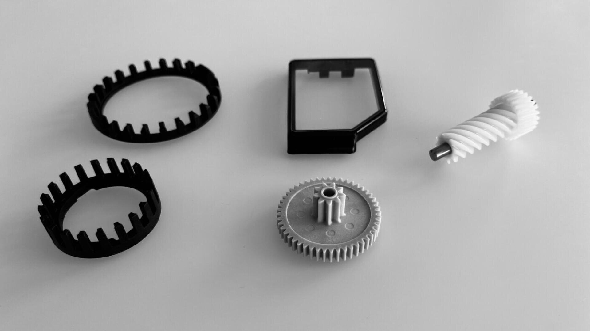 Five mechanical parts are arranged on a white surface, including two black plastic rings, a rectangular clear case, a white gear with a vertical rod, and a white helical gear. The image is in black and white.