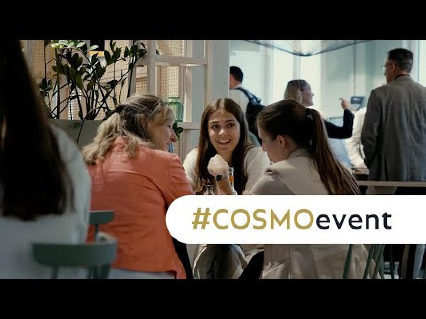 Drei Frauen sitzen an einem Tisch und unterhalten sich bei einer belebten Veranstaltung in einer Halle, mit anderen Personen im Hintergrund. Ein Banner mit #COSMOevent ist über den unteren Teil des Bildes gelegt.