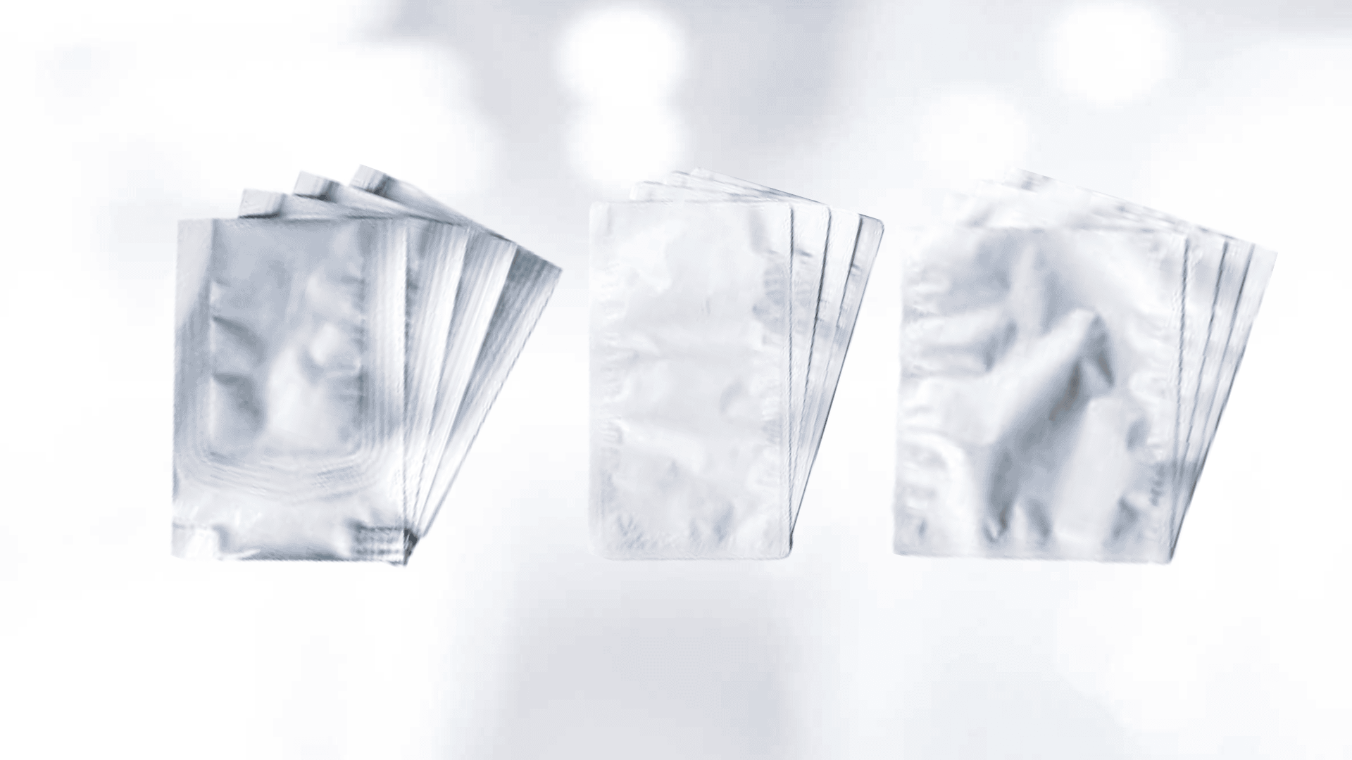 Three groups of shiny, silver foil packets are arranged against a soft, light blue, blurred background. Each group shows several sealed rectangular packets stacked slightly overlapping.