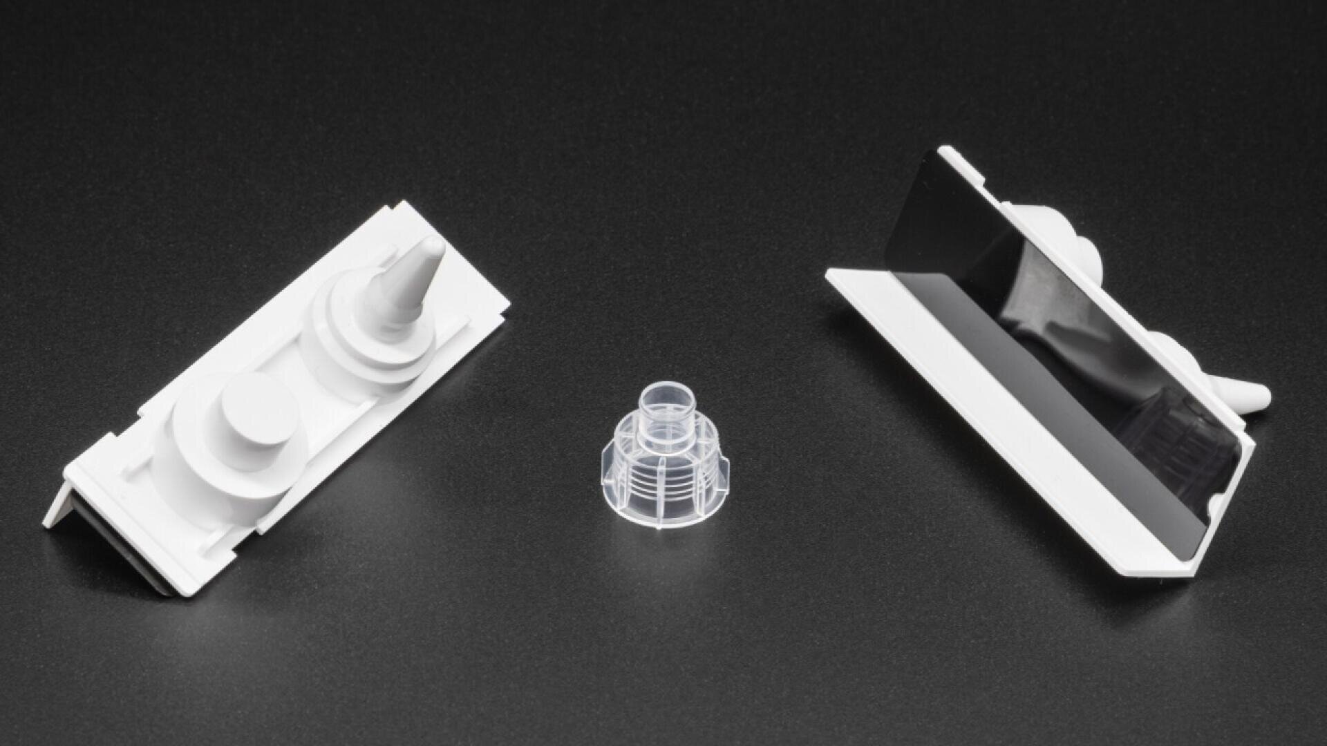 Three small plastic components are arranged on a black surface: two white parts with geometric shapes on either side, and a clear, cylindrical, ribbed piece in the center.