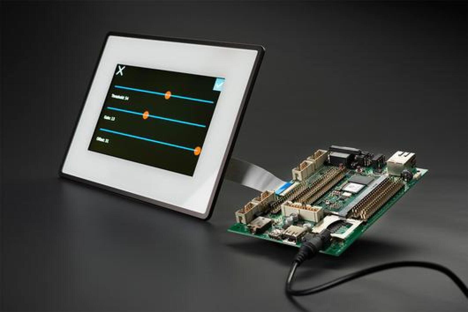 A touchscreen display with a control interface is connected via a ribbon cable to a green circuit board with connectors and components on a dark background.