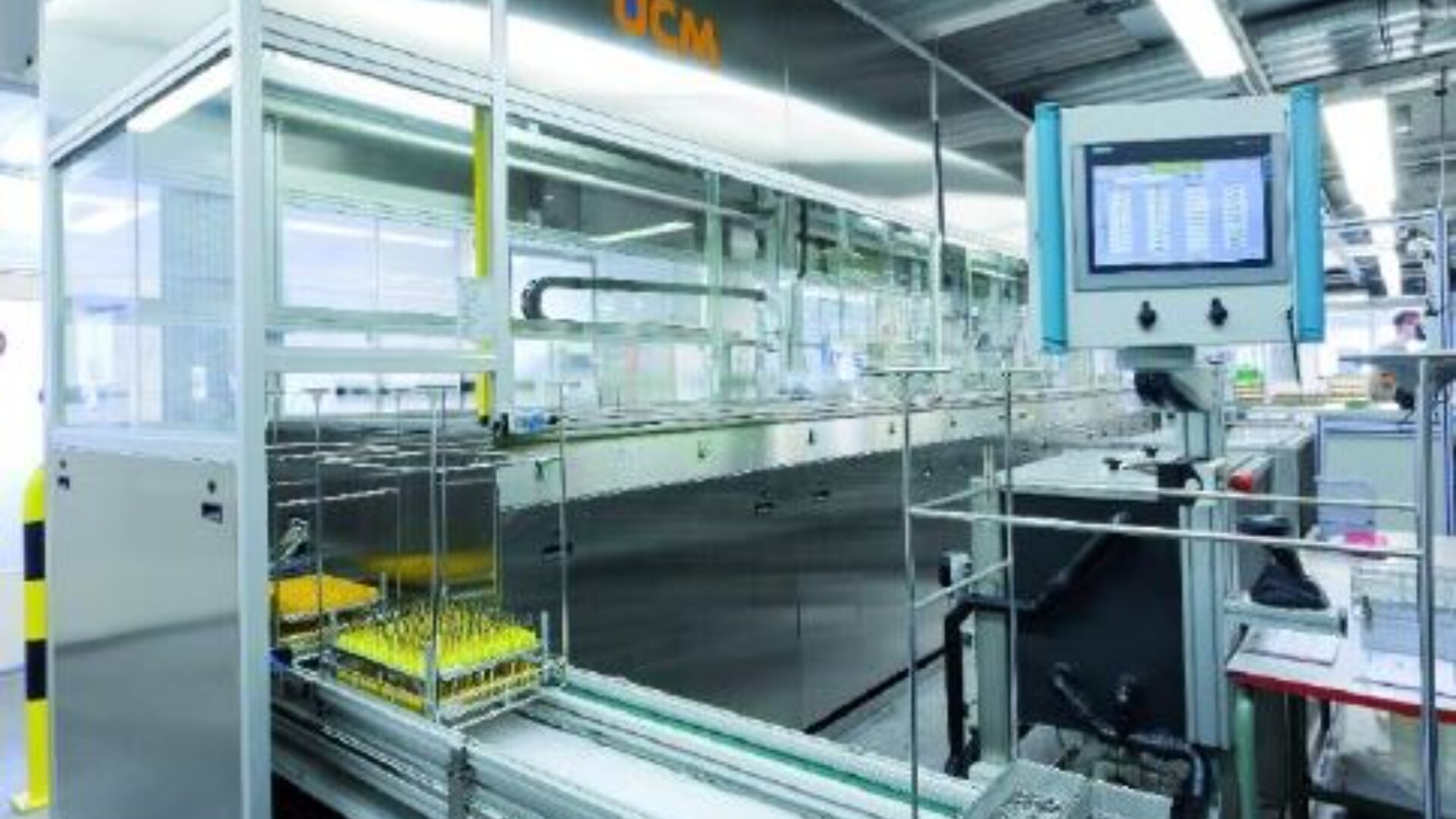 Eine moderne Industriemaschine mit einem Bedienfeld und einer Glasabdeckung verarbeitet Tabletts mit Artikeln in einer hell erleuchteten, automatisierten Fabrikumgebung.