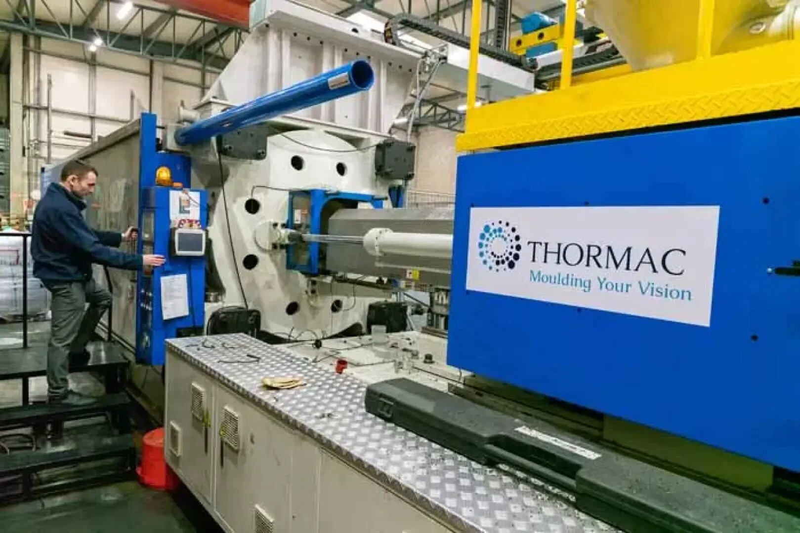 A person operates a large industrial injection molding machine inside a factory. The machine displays a label with the company name Thormac and the slogan Moulding Your Vision.”.