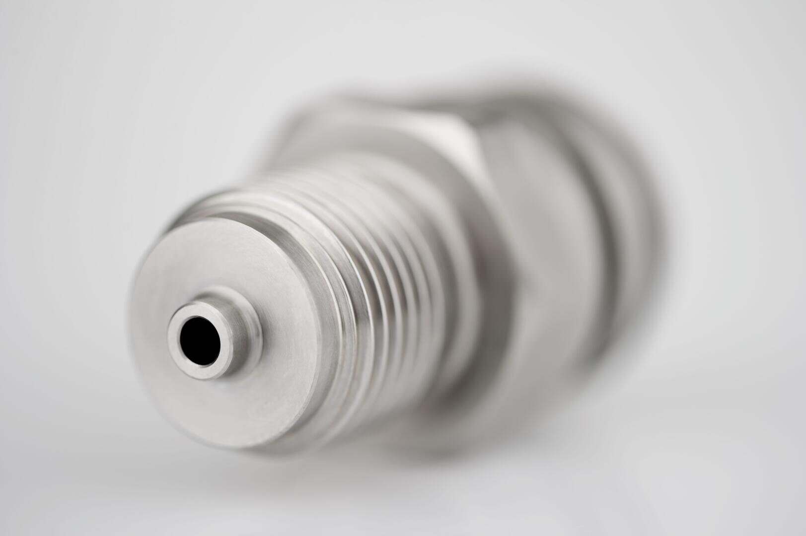 Nahaufnahme einer metallischen Zündkerze vor unscharfem Hintergrund, mit Fokus auf Gewinde und Spitze. Das Bild hebt die glatte, glänzende Oberfläche und die präzisen technischen Details des Teils hervor.