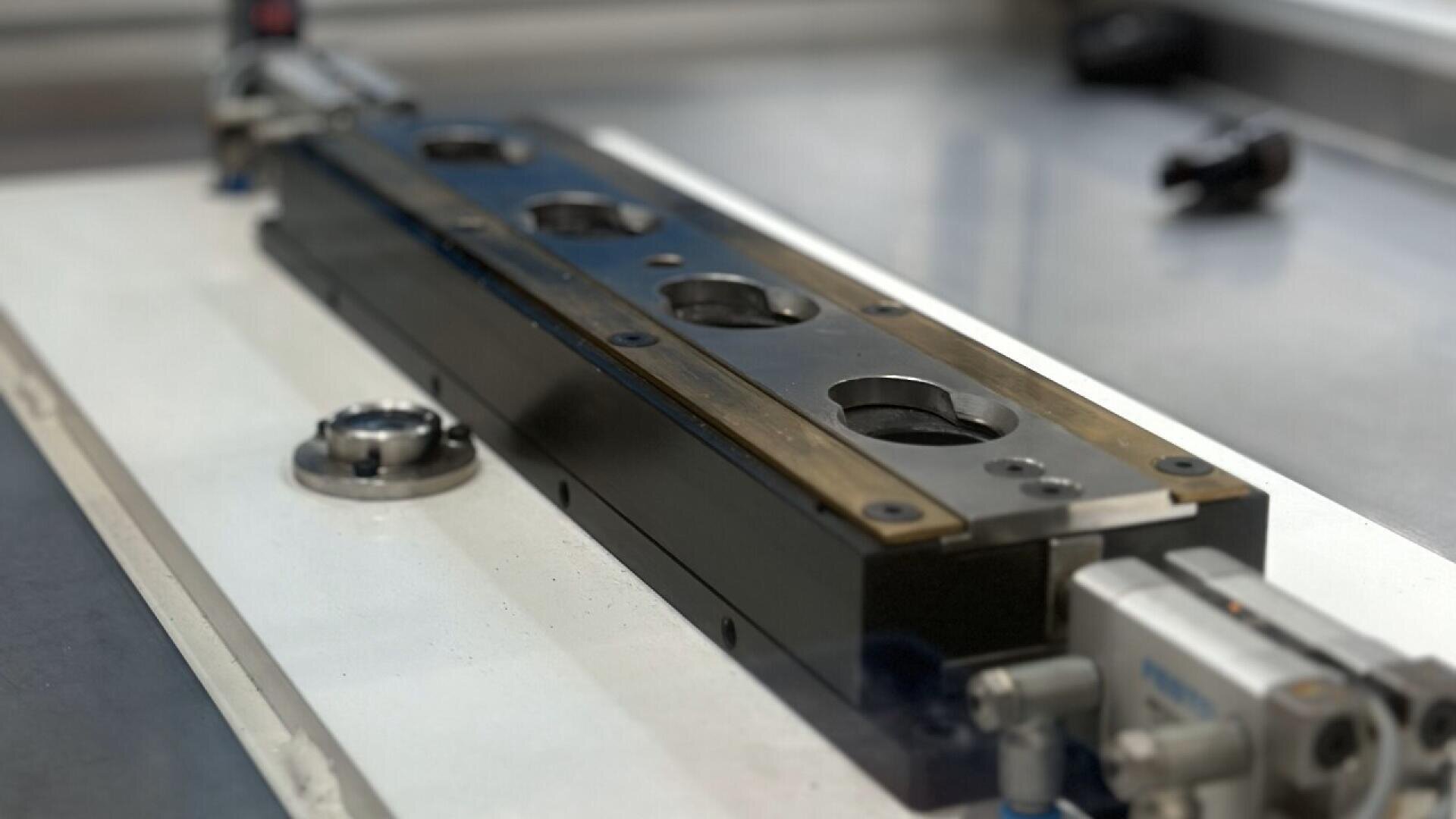 A close-up of a precision metal machine component with circular cutouts, mounted on a white work surface in a workshop or laboratory setting.
