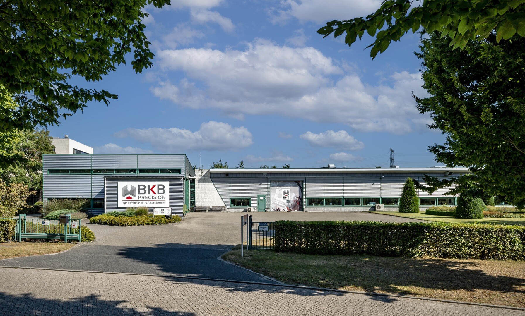Modernes Industriegebäude mit BKB Precision-Beschilderung an der Vorderseite, umgeben von grünen Rasenflächen, Bäumen und einer gepflasterten Auffahrt unter einem teilweise bewölkten blauen Himmel.