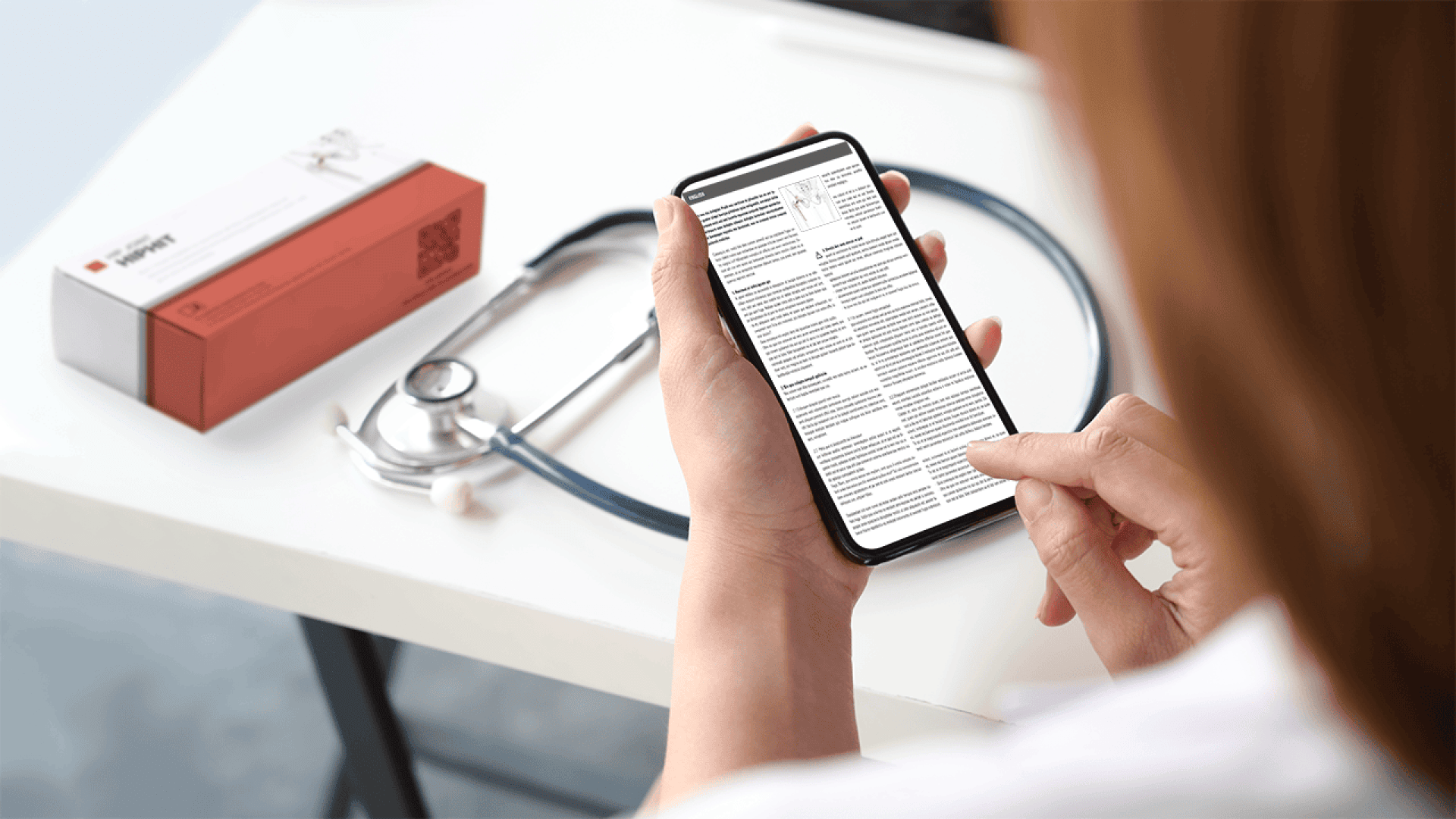 A person in a white coat uses a smartphone to read a document. A stethoscope and a medicine box are on the table beside them.