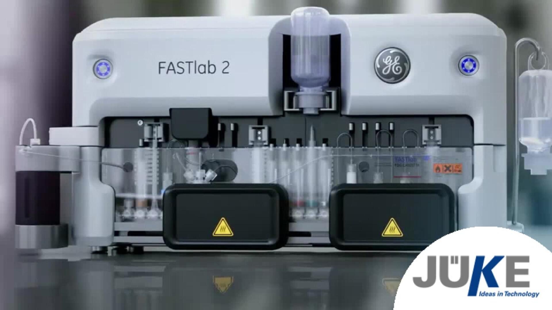 A GE FASTlab 2 laboratory instrument with clear compartments, warning symbols, and visible internal components; the bottom right features a JUKE logo with the tagline Ideas in Technology.