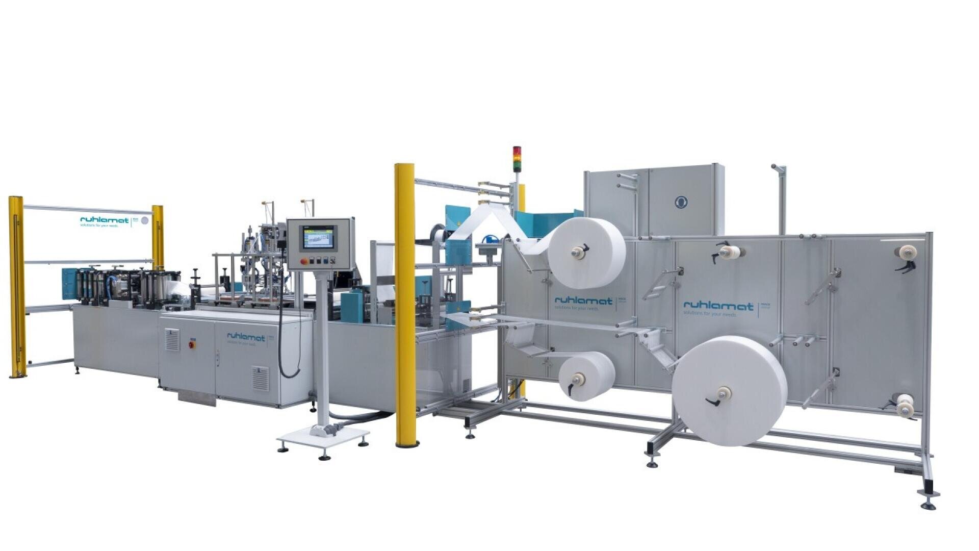 Industrial mask production machine with control panel, yellow safety poles, and multiple large white rolls of material set in a clean, white environment. The machine is labeled “ruhlamat.”.