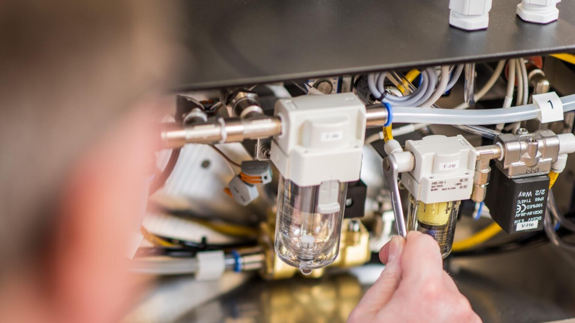 Die Hand einer Person benutzt ein Werkzeug, um Komponenten in einer Maschine einzustellen oder zu reparieren. Dabei werden verschiedene Drähte, Schläuche und Industrieteile miteinander verbunden. Das Bild konzentriert sich auf die mechanischen Komponenten.