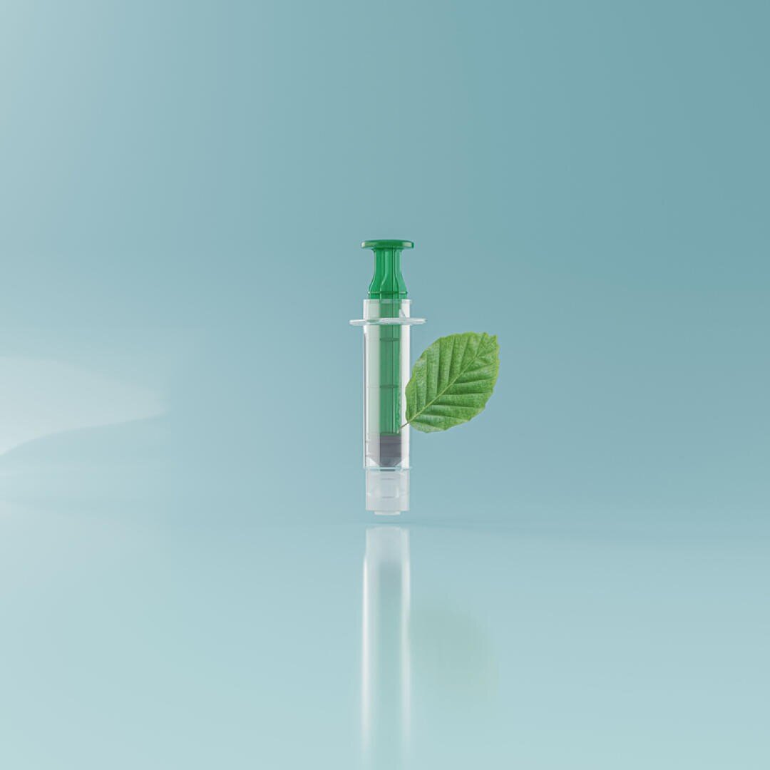 A syringe filled with green liquid stands upright on a light blue background. A green leaf is attached to its side, symbolizing environmentally friendly or natural medical solutions.