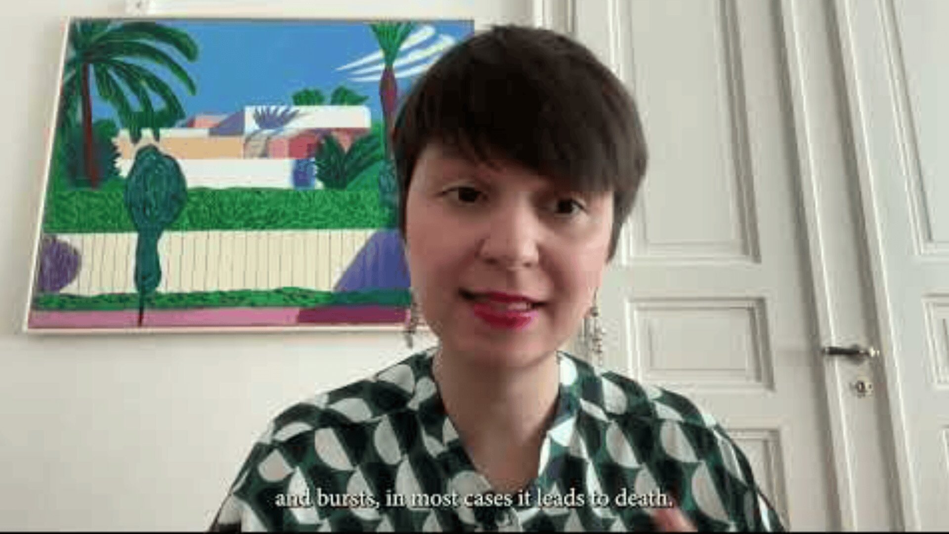 A woman with short dark hair and red lipstick speaks on camera in a bright room with a colorful painting and white double doors behind her. Subtitles read: “and bursts, in most cases it leads to death.”.