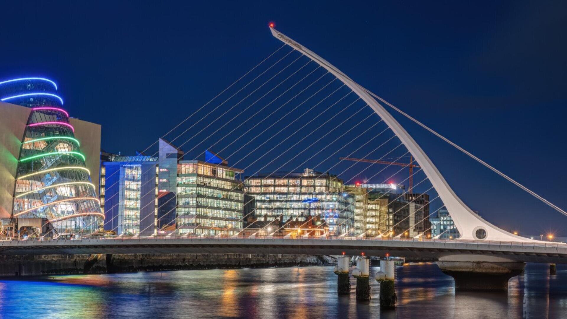 A modern cable-stayed bridge lit at night spans a river, with colorful illuminated buildings and glass offices in the background, reflecting on the water.