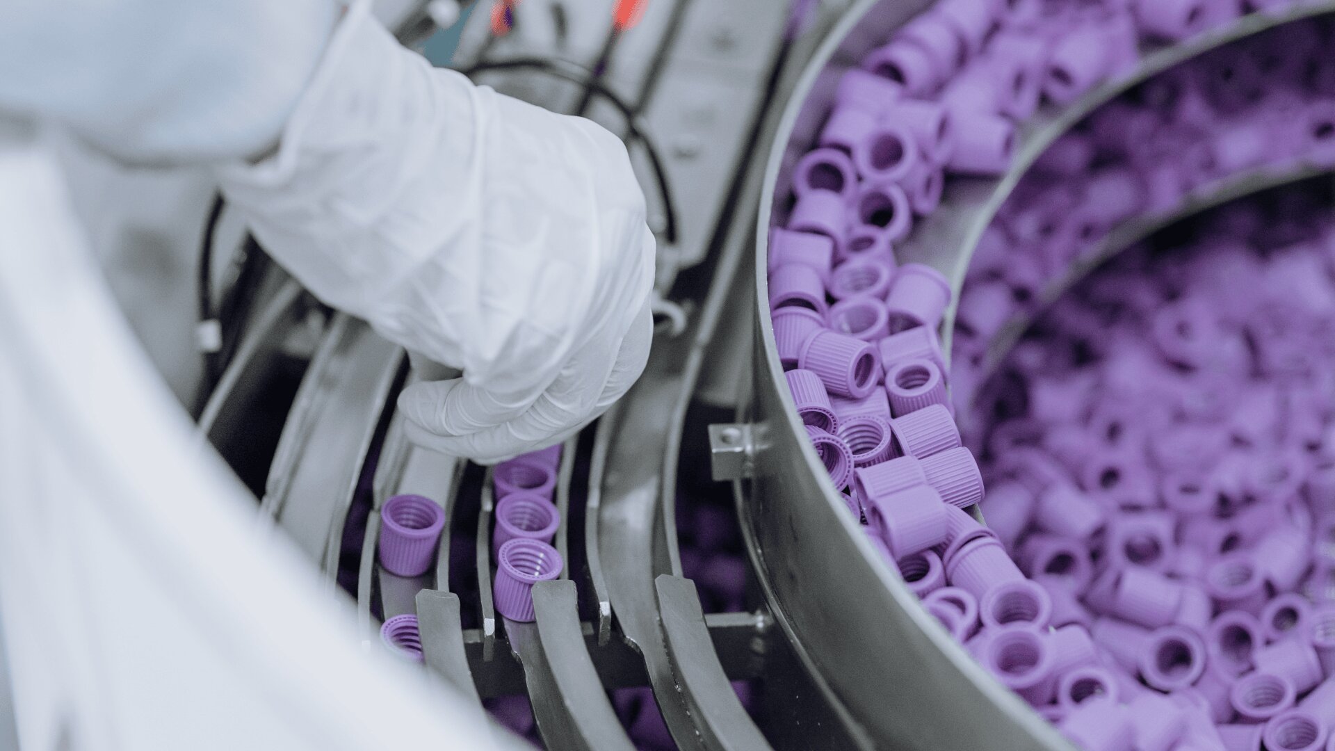 A gloved hand operates machinery sorting numerous purple plastic caps in a production or laboratory setting.