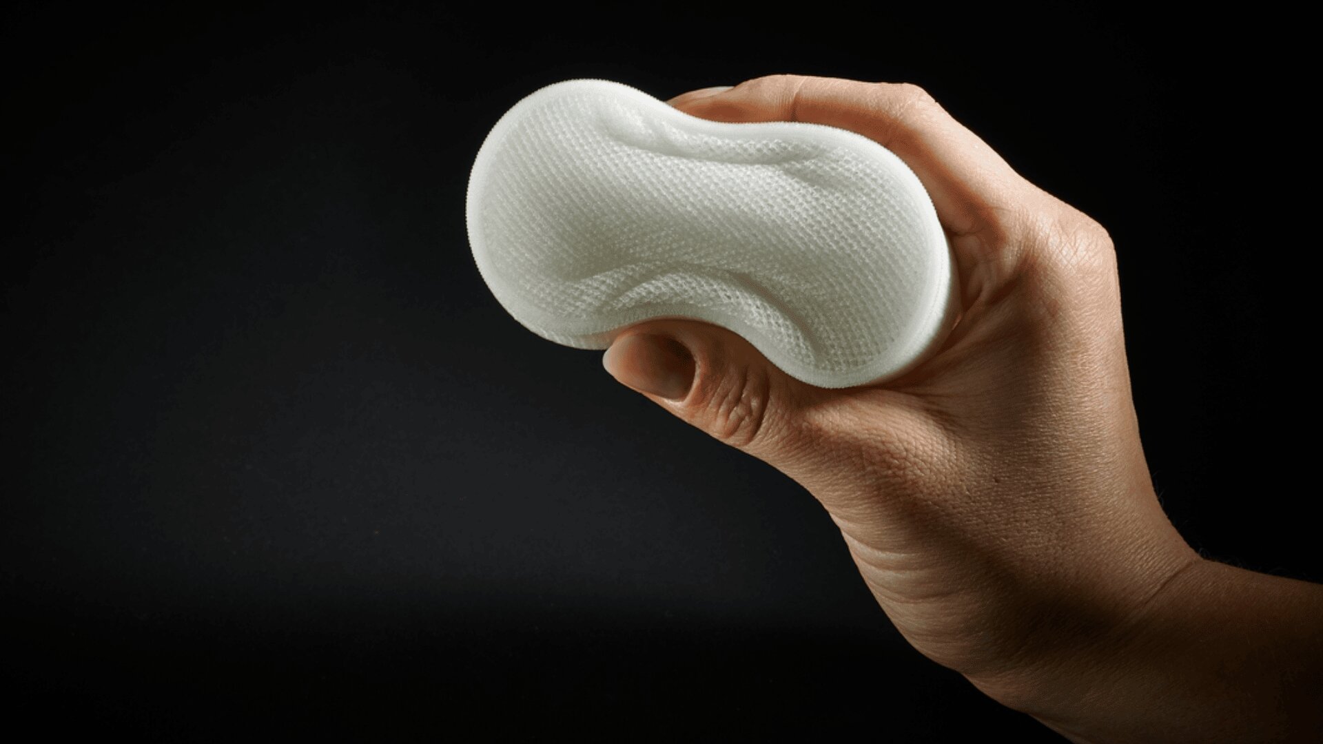 Eine Hand hält ein weißes, strukturiertes und gebogenes Objekt vor schwarzem Hintergrund. Das Objekt wirkt weich und flexibel, mit einer welligen Oberfläche und Konturen, die sich der Form der Hand anpassen.