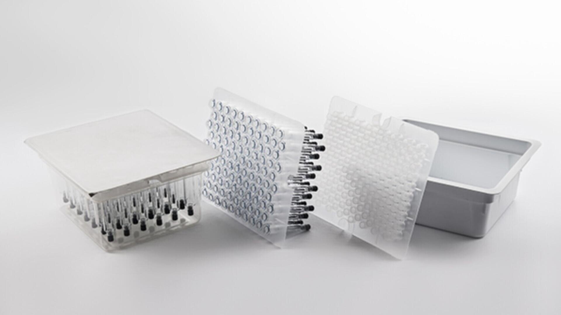A packaging tray filled with small glass vials, a plastic cover, a transparent molded tray with more vials, and a gray box are arranged on a white surface.