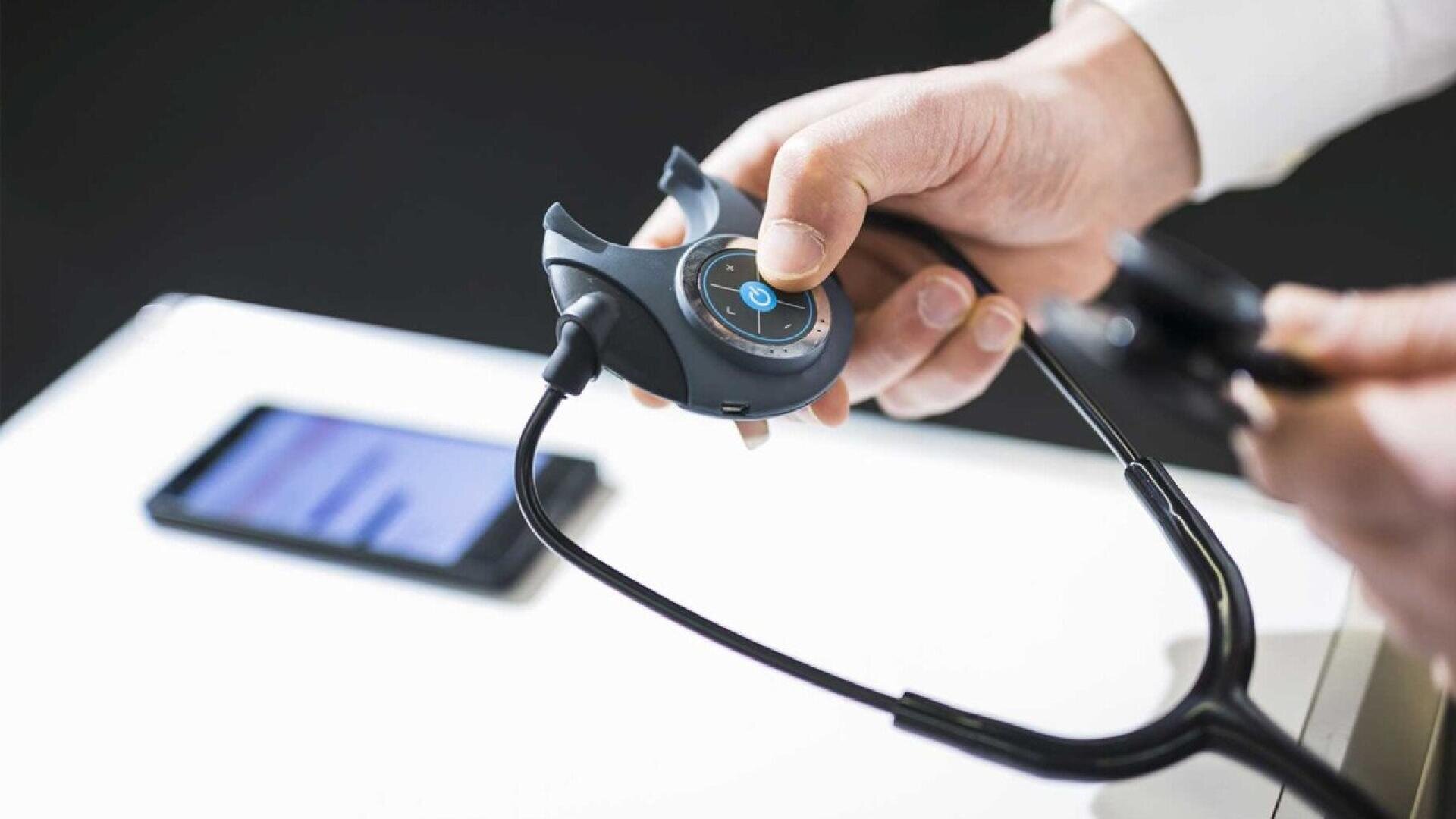 A person holds a modern digital stethoscope, adjusting its controls. A smartphone with a blurred screen is on the table in the background, suggesting the use of technology in healthcare.