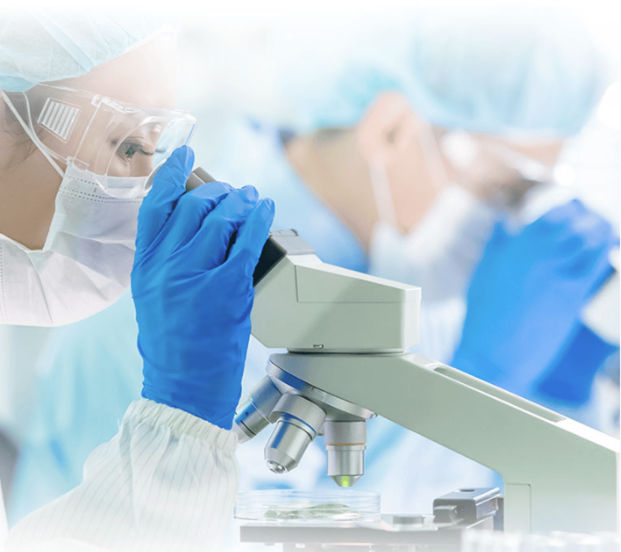 A scientist in protective clothing, gloves, and safety goggles looks through a microscope in a laboratory while another scientist works in the background.