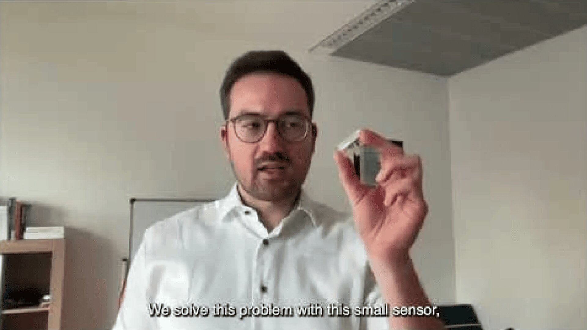 A man in glasses and a white shirt holds up a small sensor device in a room. Subtitles at the bottom read, We solve this problem with this small sensor,.