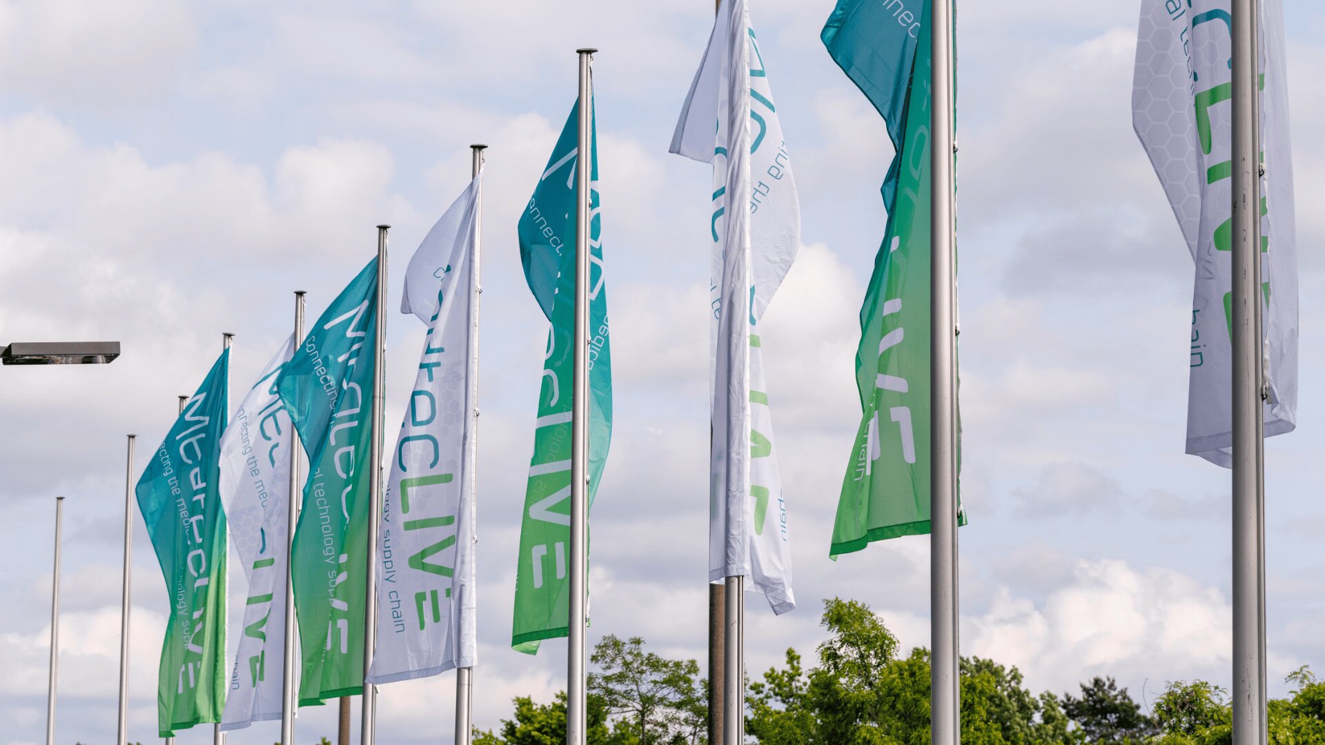 A row of teal and white flags with medLIVE and other text wave on flagpoles outdoors against a cloudy sky, with some trees visible in the background.