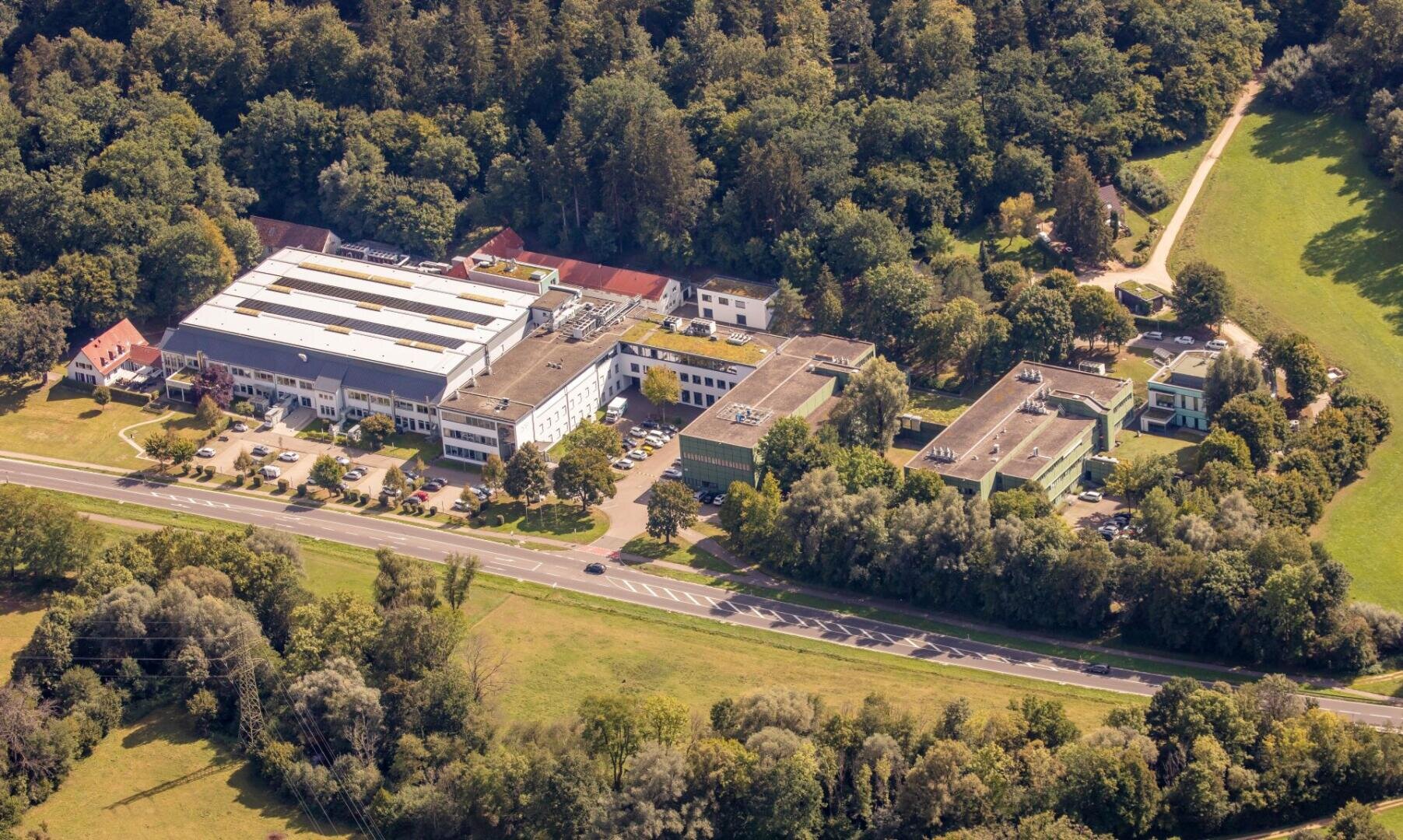 Aerial view of a business or industrial complex surrounded by trees, situated next to a main road with smaller buildings, parking lots, and a forested area in the background.