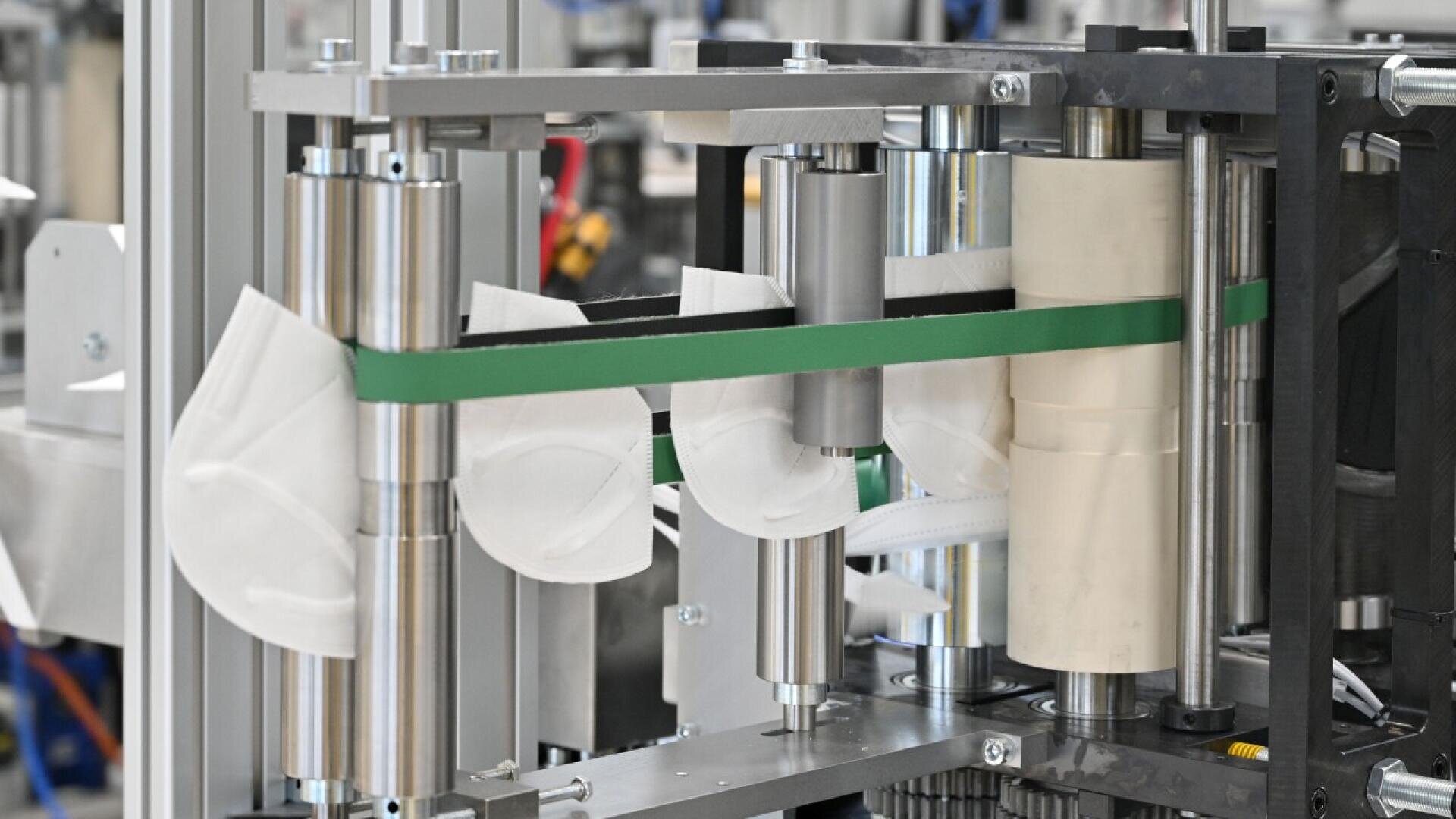 A close-up of a machine in a factory assembling white face masks, showing rollers, a green conveyor belt, and partially completed masks in the production process.