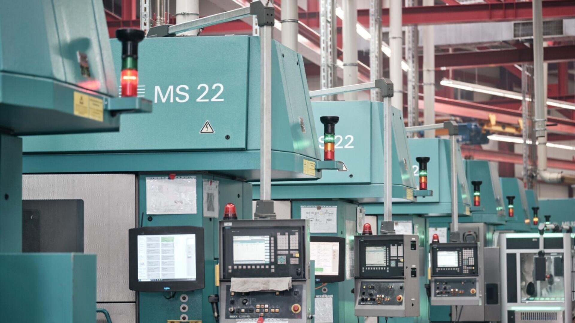 A row of teal industrial machines with control panels and indicator lights in a modern factory setting, with red pipes and overhead lights visible in the background.
