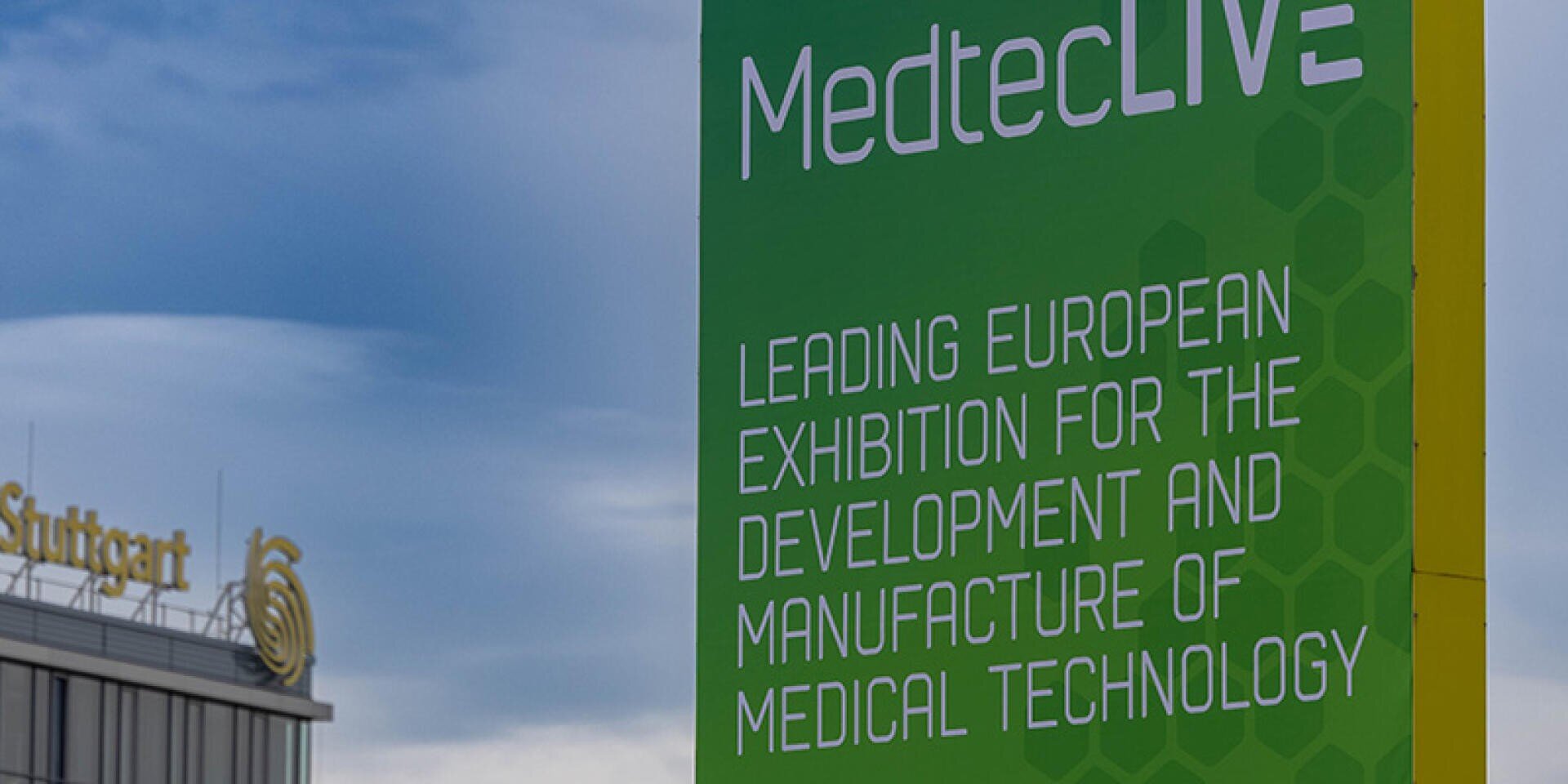 Auf einem grünen Schild steht „MedtecLIVE: Europas führende Fachmesse für die Entwicklung und Herstellung von Medizintechnik“. Im Hintergrund ist unter einem bewölkten Himmel ein Gebäude in Stuttgart zu sehen.