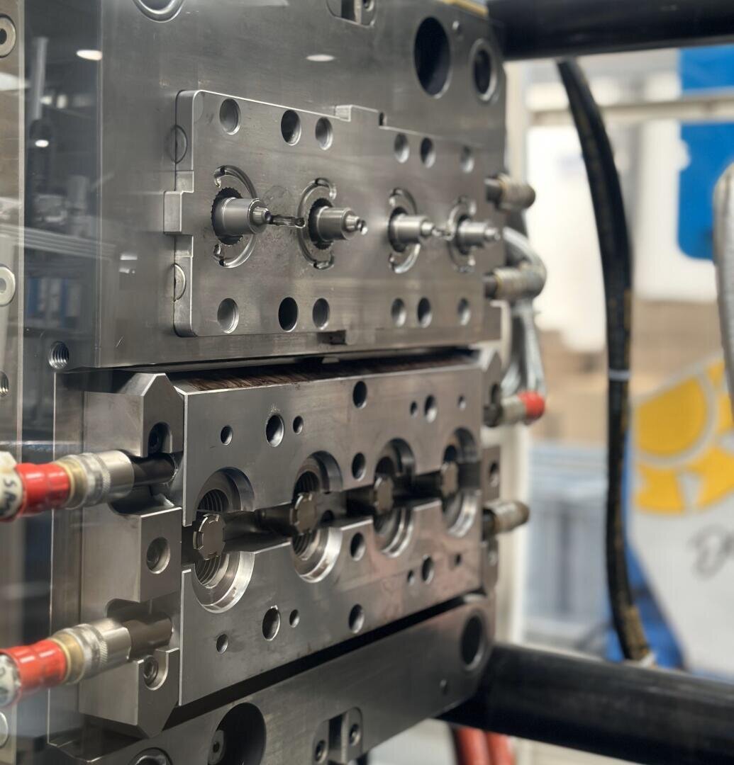 Nahaufnahme einer Metallspritzgussform in einer Fertigungsmaschine. Sie zeigt detaillierte Hohlräume und Kanäle, mit roten und schwarzen Rohren, die mit der Form verbunden sind, und einer industriellen Umgebung im Hintergrund.