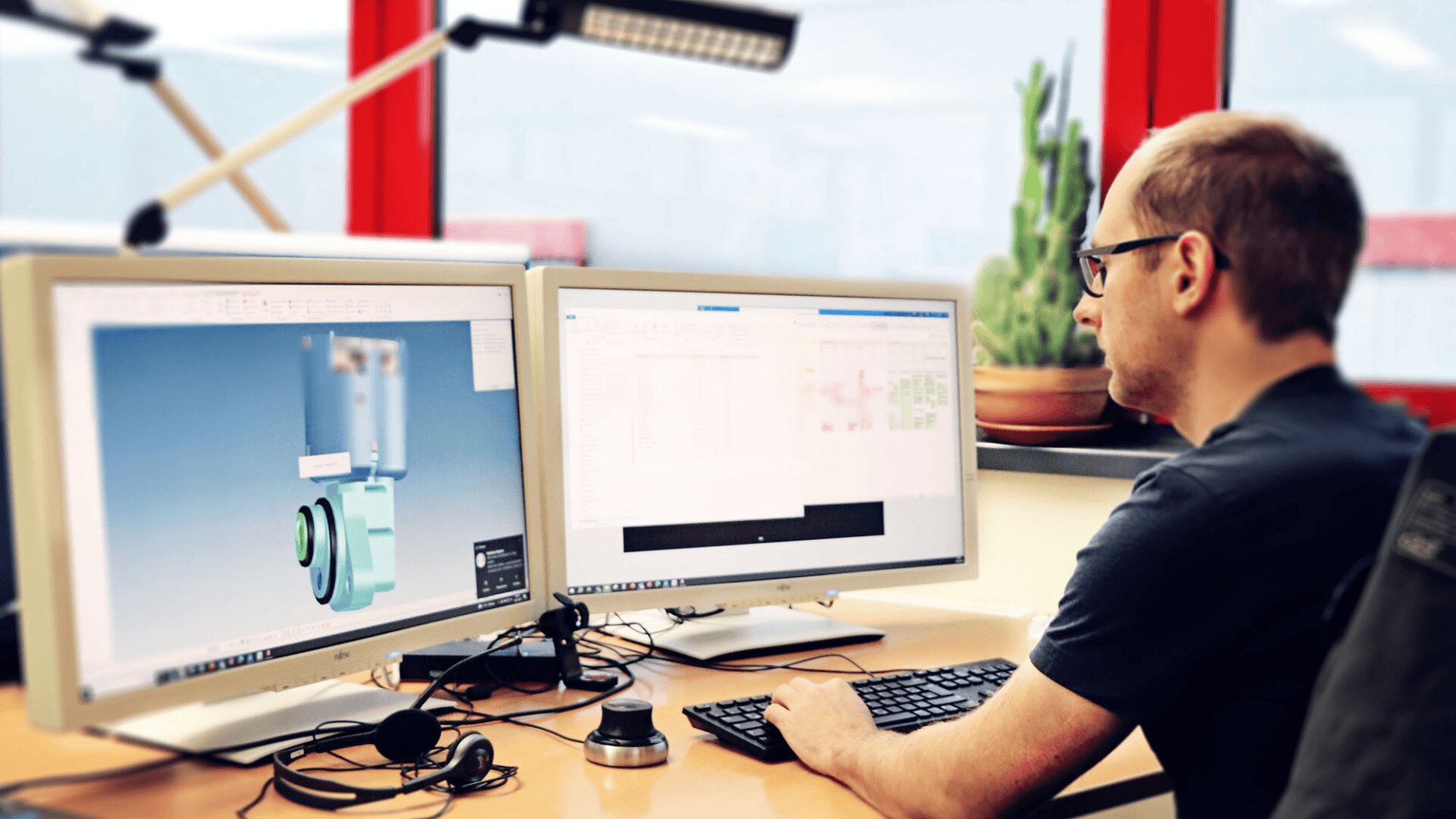 Eine Person sitzt an einem Schreibtisch und arbeitet an zwei Computermonitoren, von denen einer ein 3D-Modell anzeigt, möglicherweise in einer Designsoftware. Der Arbeitsbereich umfasst eine Tastatur, eine Maus, eine Pflanze und natürliches Licht durch große Fenster.
