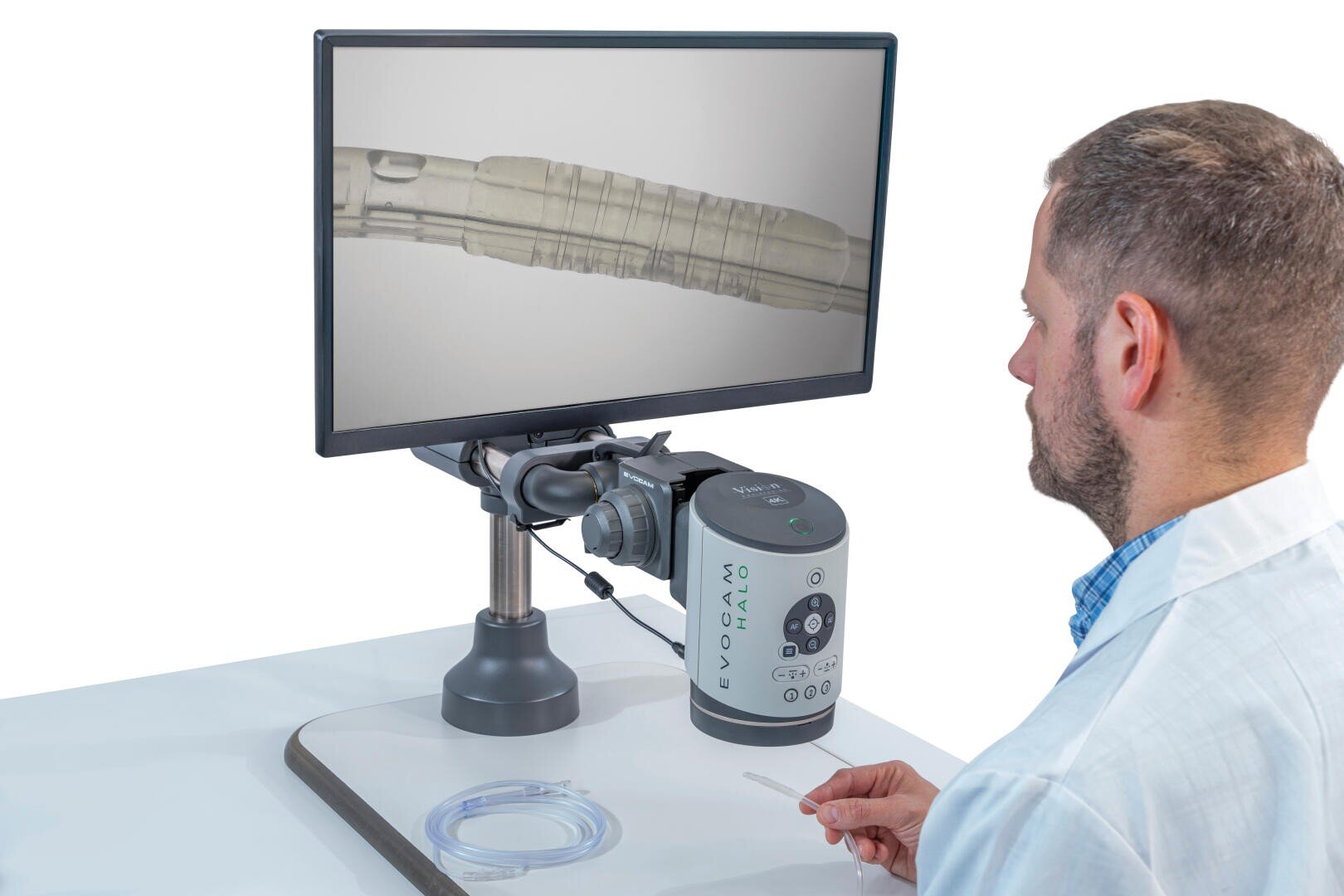 Ein Mann im Laborkittel untersucht eine Probe mit einem Digitalmikroskop, das an einen Monitor angeschlossen ist, der ein vergrößertes Bild eines transparenten Röhrchens oder einer biologischen Probe anzeigt. Auf dem weißen Tisch sind Laborgeräte angeordnet.