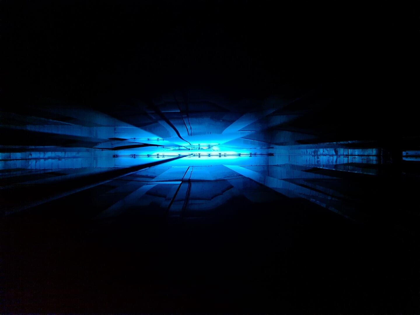 Abstraktes Bild, das einen dunklen Raum mit einem hellen blauen Licht in der Mitte zeigt, das Schlieren und Reflexionen erzeugt, die nach außen strahlen und ein Gefühl von Tiefe und Symmetrie vermitteln.