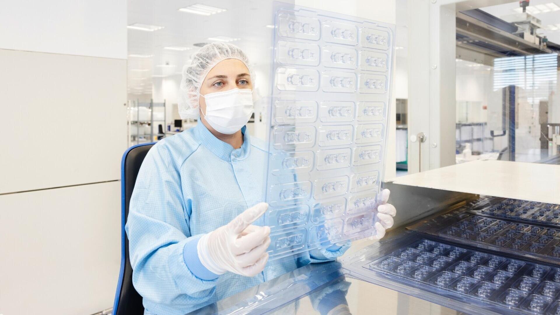 A person in protective clothing, including a hair net, mask, gloves, and a blue lab coat, examines a clear plastic tray with multiple compartments in a clean, bright laboratory setting.