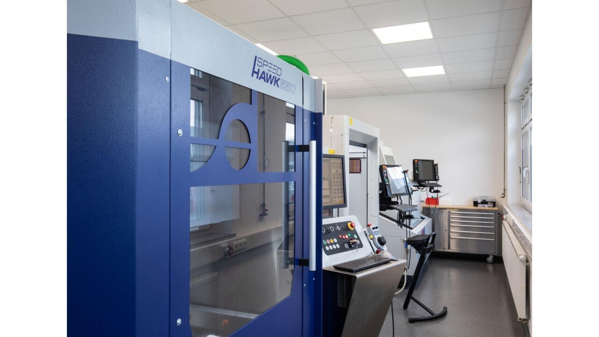 A modern laboratory or manufacturing room with blue and white ISPER HAWK 650 machinery, control panels, monitors, and bright overhead lighting.