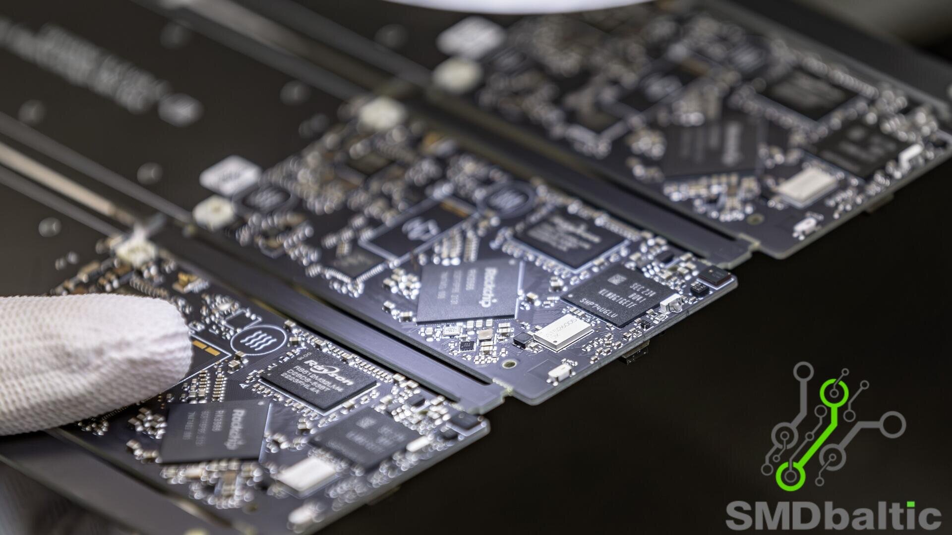 Eine behandschuhte Hand hält eine schwarze Leiterplatte (PCB) mit verschiedenen Mikrochips und elektronischen Komponenten neben anderen PCBs. In der unteren rechten Ecke ist das Logo von SMD Baltic zu sehen.