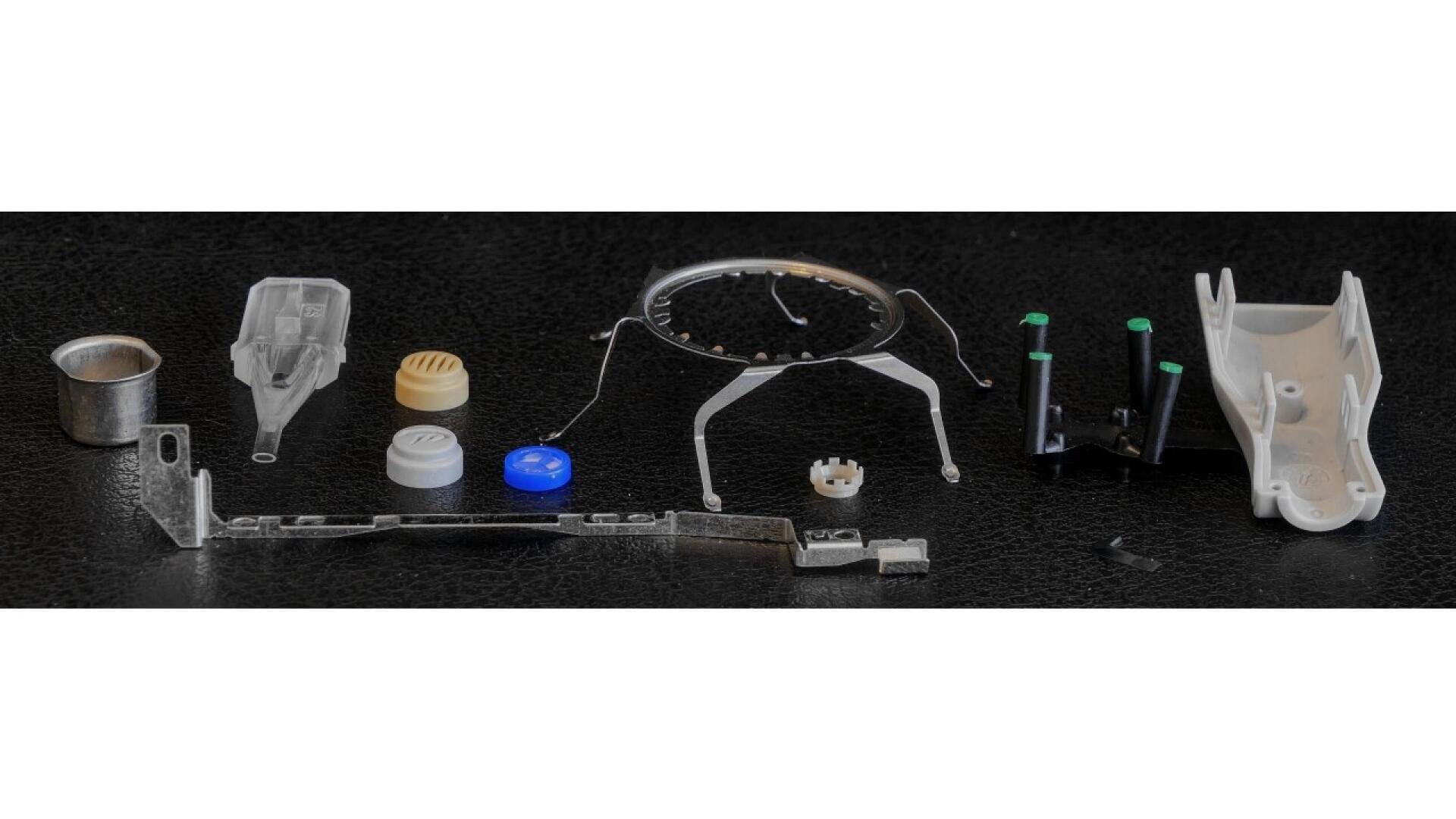 Assorted small mechanical and plastic components, including gears, clips, and brackets, are neatly arranged on a dark surface, showcasing a variety of shapes, colors, and materials.