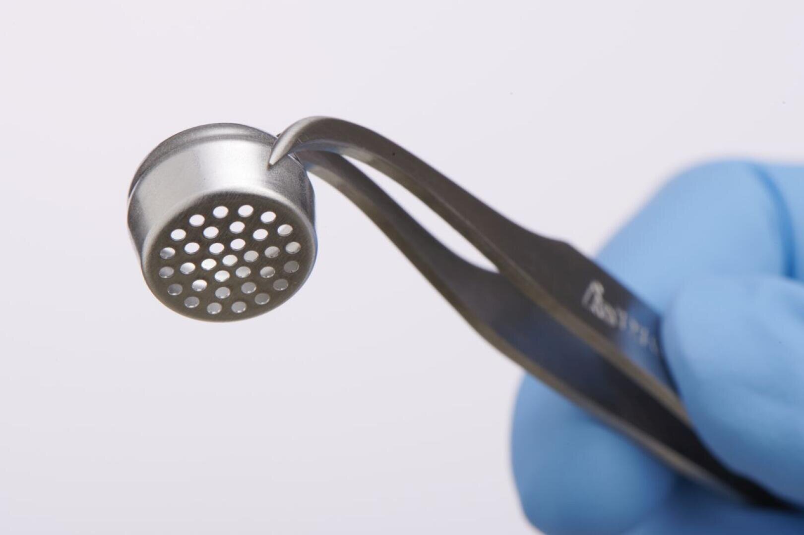 A gloved hand uses tweezers to hold a small, round metal object with multiple holes on one side against a plain, light background.