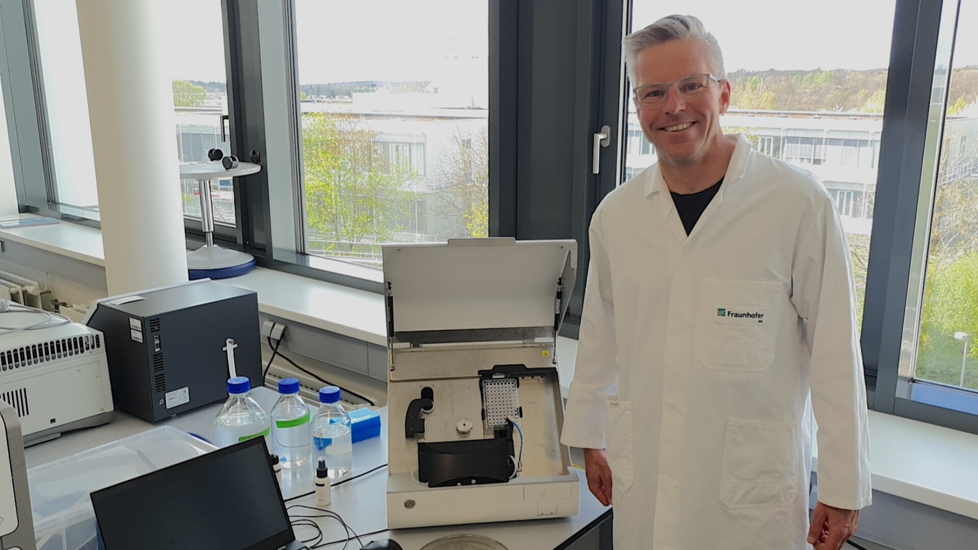 Andreas Traube im weißen Laborkittel steht neben offenen Laborgeräten auf einem Tisch mit Flaschen und einem Laptop in einem hellen Labor mit großen Fenstern und malerischer Aussicht nach draußen.
