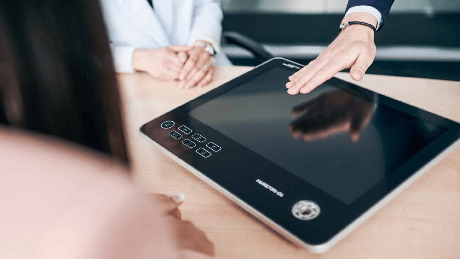 Eine Nahaufnahme von zwei Personen, die an einem Schreibtisch sitzen, wobei eine Person auf ein großes Touchscreen-Gerät mit einem leeren Bildschirm zeigt oder darüber gestikuliert.