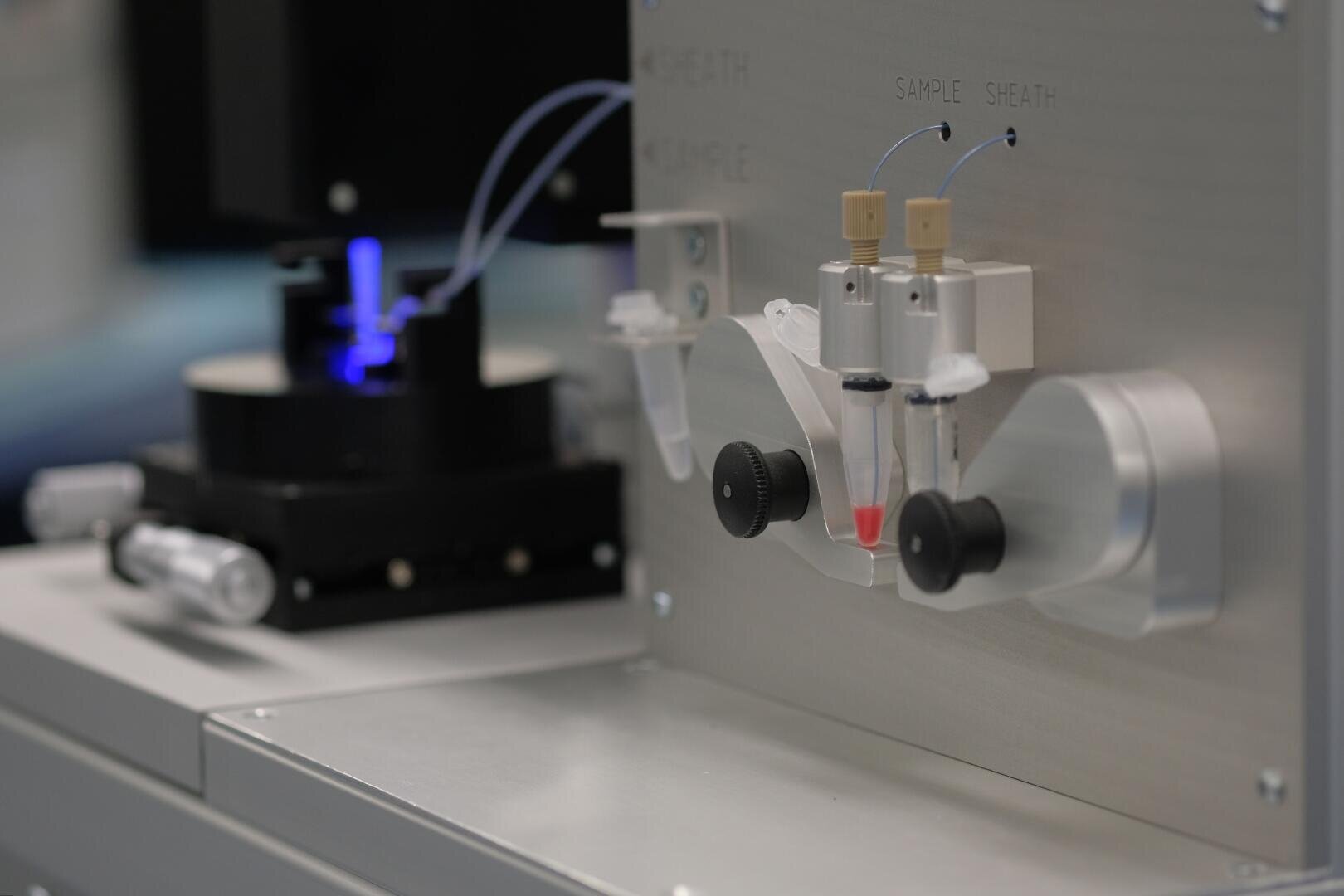 Nahaufnahme eines Laborgeräts mit beschrifteten Anschlüssen für Proben und Ummantelung, Röhrchen, die an Fläschchen angeschlossen sind, und einem blauen Licht im Hintergrund, das für wissenschaftliche oder medizinische Analysen verwendet wird.