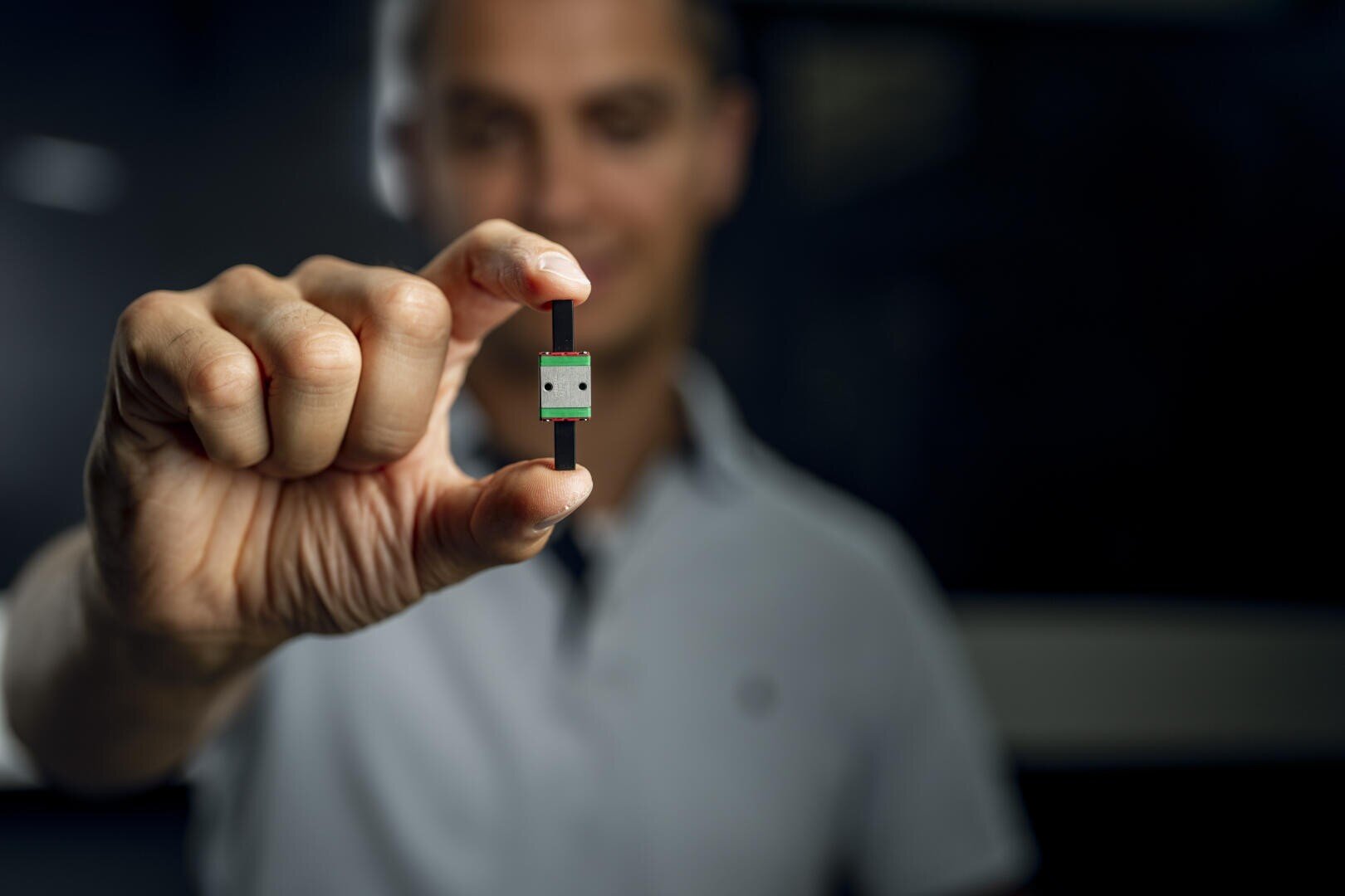 A person in a light-colored shirt holds a small, rectangular electronic component between thumb and forefinger, with the background gently blurred.